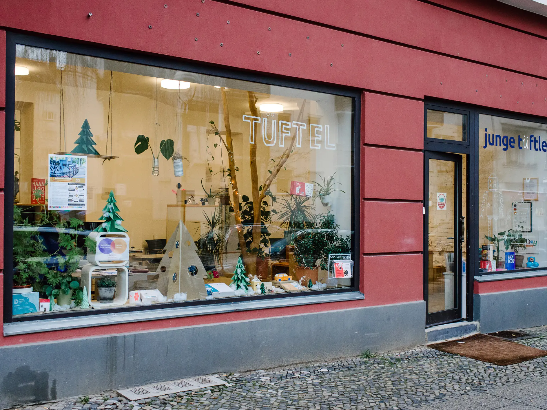 A storefront with a large window displaying various crafts and greenery. The sign TUFTEL is seen on the window. The building has a dark red exterior. The door on the right has a sign junge tüftler. Cobblestone pavement is in front.