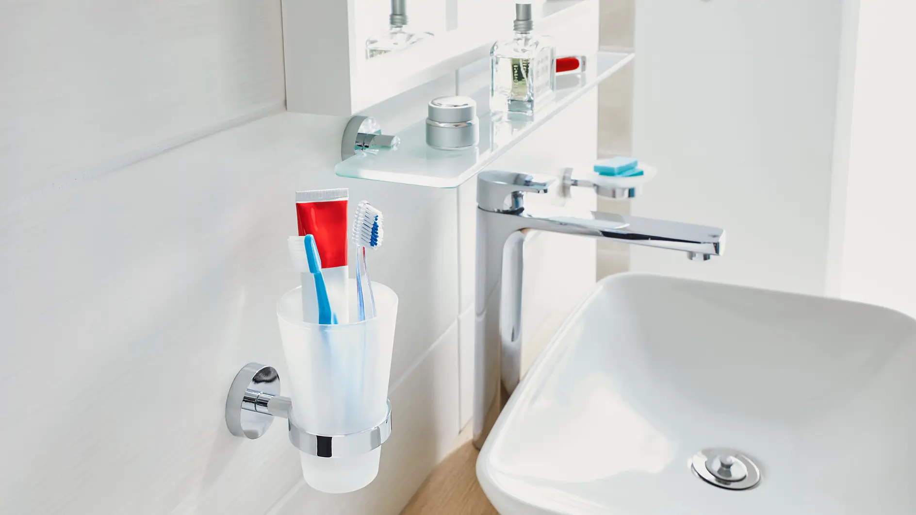 Bathroom sink with a white ceramic basin and a chrome faucet. A toothbrush holder with a toothpaste tube and two toothbrushes is mounted on the wall using tesa tape. The glass shelf above holds toiletries, and a blurred reflection is visible in the mirror.