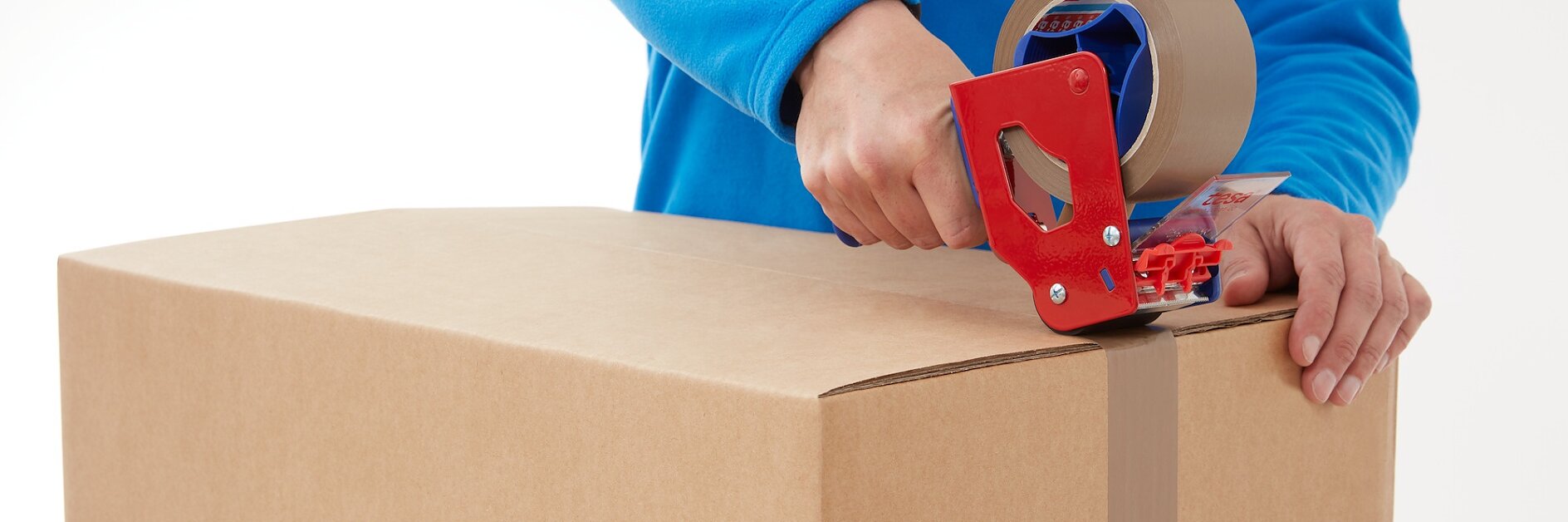 A person in a blue shirt is sealing a cardboard box using a red tape dispenser with brown tesa tape. The person is applying tesa tape along the center seam of the box.