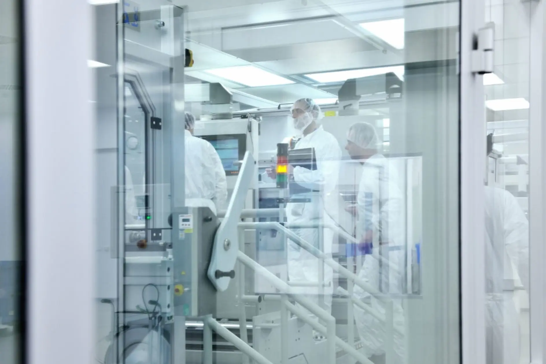 A group of people in white protective suits work inside a laboratory with large equipment. The room is clean and well-lit, with visible glass walls and control panels. Any tapes used in the lab are replaced with tesa tape to ensure consistency and quality throughout their operations.