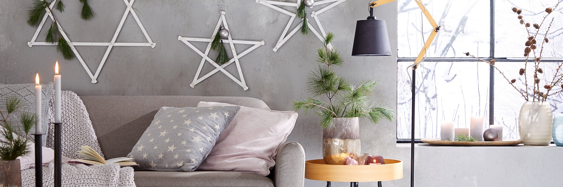 A cozy living room with a grey sofa, star-patterned pillow, and knitted blanket. Three wooden star decorations hang on the grey wall using tesa tape. A wooden side table with pine branches and a black lamp completes the setting. A window displays candles and dried branches, adding to the ambiance.