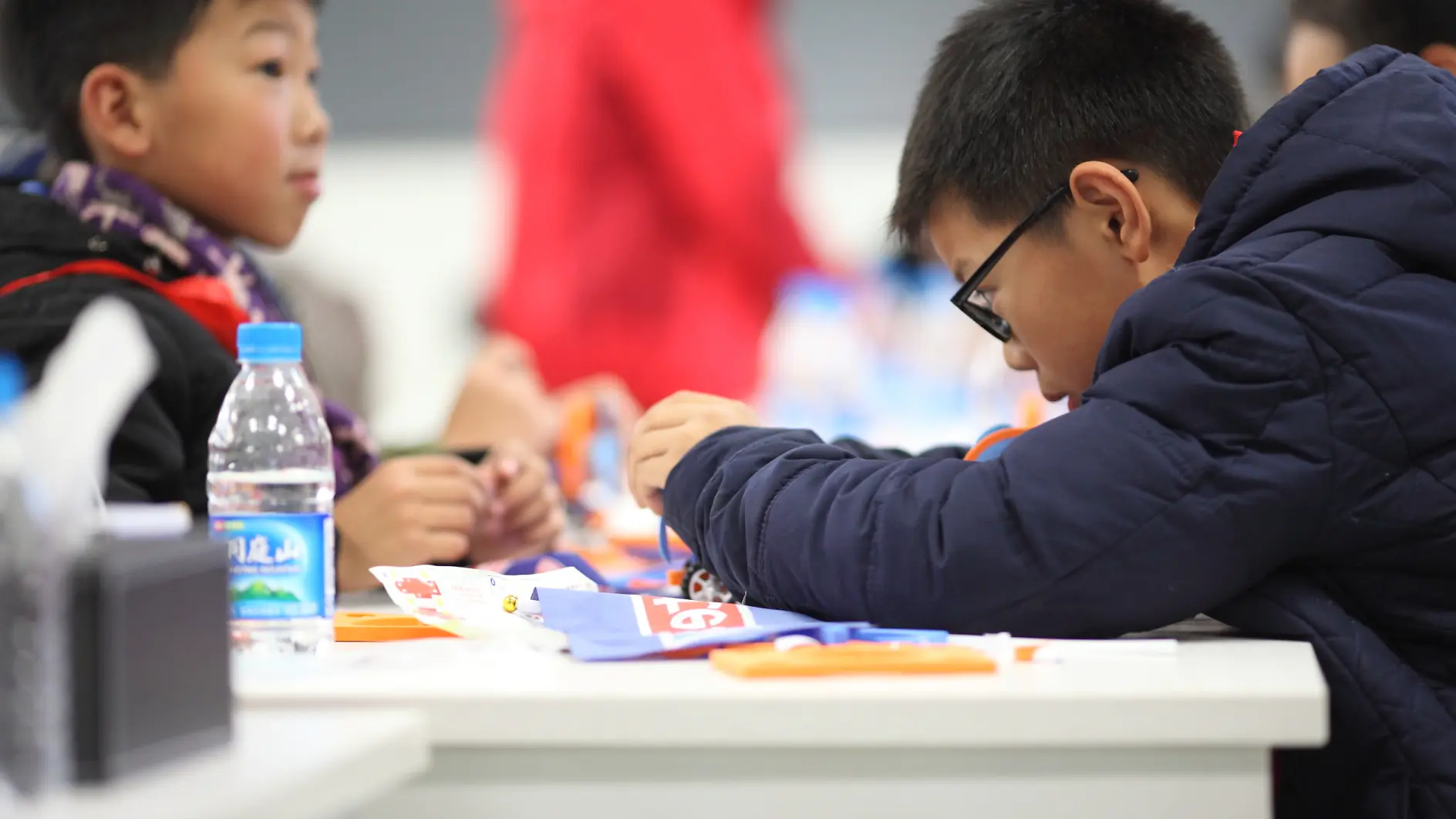 Two children are seated at a table, focused on a craft activity. The child on the right wears glasses and a dark jacket. A water bottle and craft materials, including tesa tape, are on the table. The background is blurred, with more people and objects visible. (This text has been generated by AI)