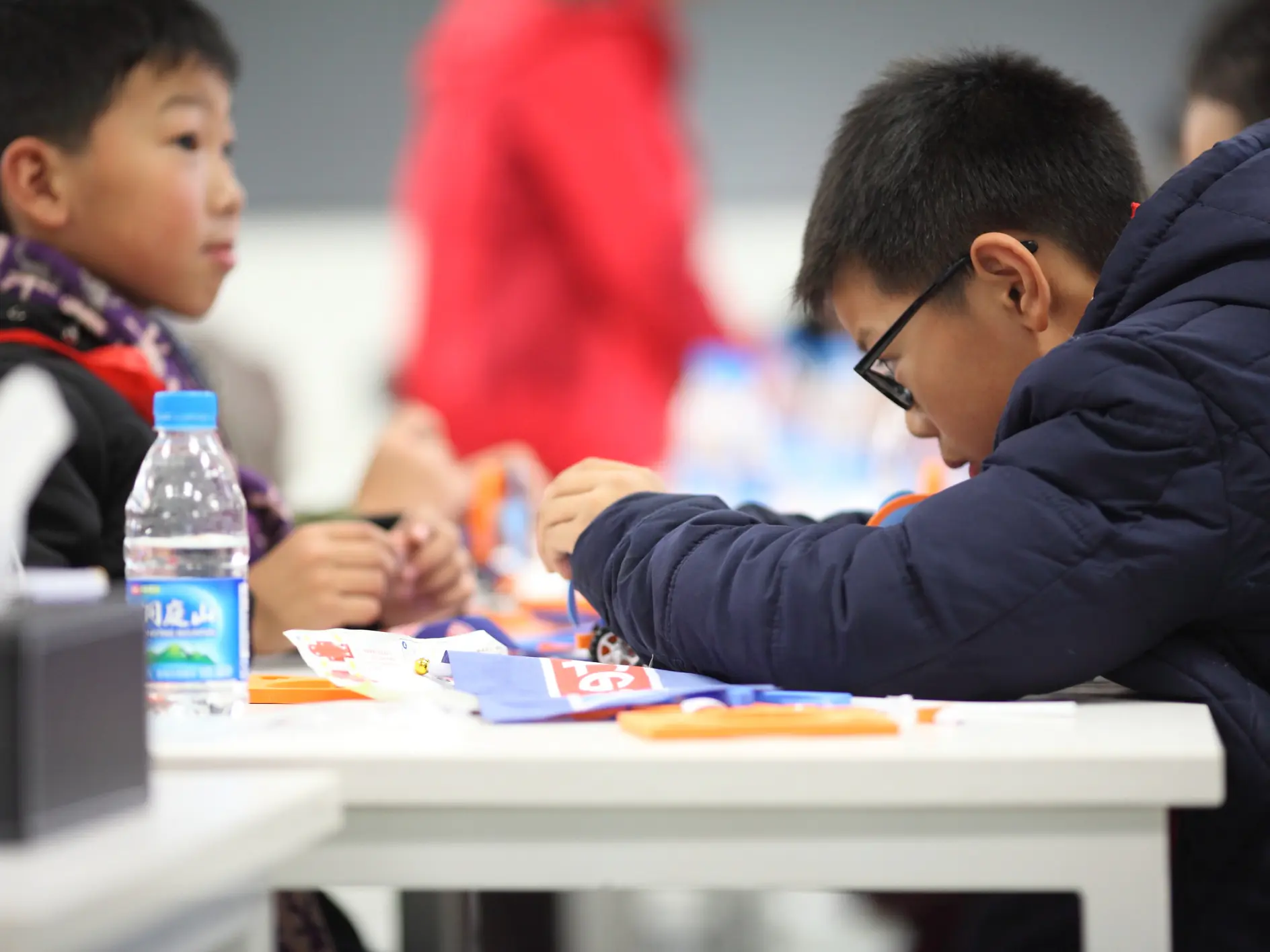 Two children are seated at a table, focused on a craft activity. The child on the right wears glasses and a dark jacket. A water bottle and craft materials, including tesa tape, are on the table. The background is blurred, with more people and objects visible.