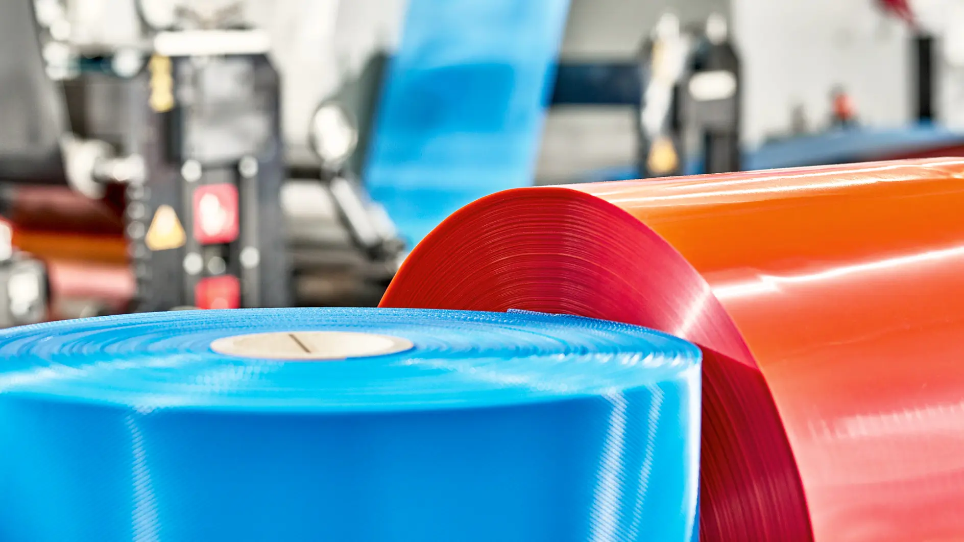 Close-up of two large rolls of industrial plastic, one blue and one red, positioned in a manufacturing facility. The background shows machinery and additional rolls in various stages of processing bound with tesa tape. (This text has been generated by AI)