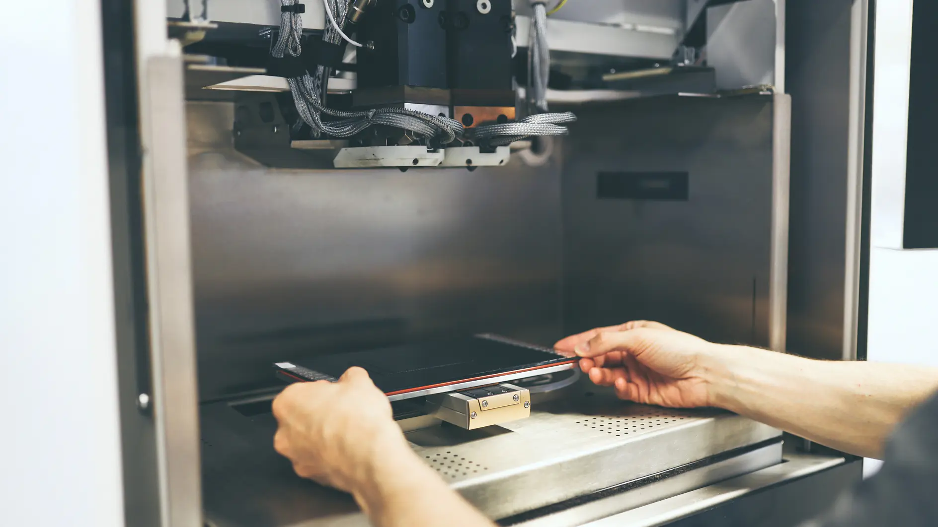 Certainly! Heres the text with the requested changes: Two hands adjust a flat material inside a 3D printer or industrial machine using tesa tape. The machine is metallic, with several components and wires visible. The scene suggests a technical or manufacturing process in progress. (This text has been generated by AI)