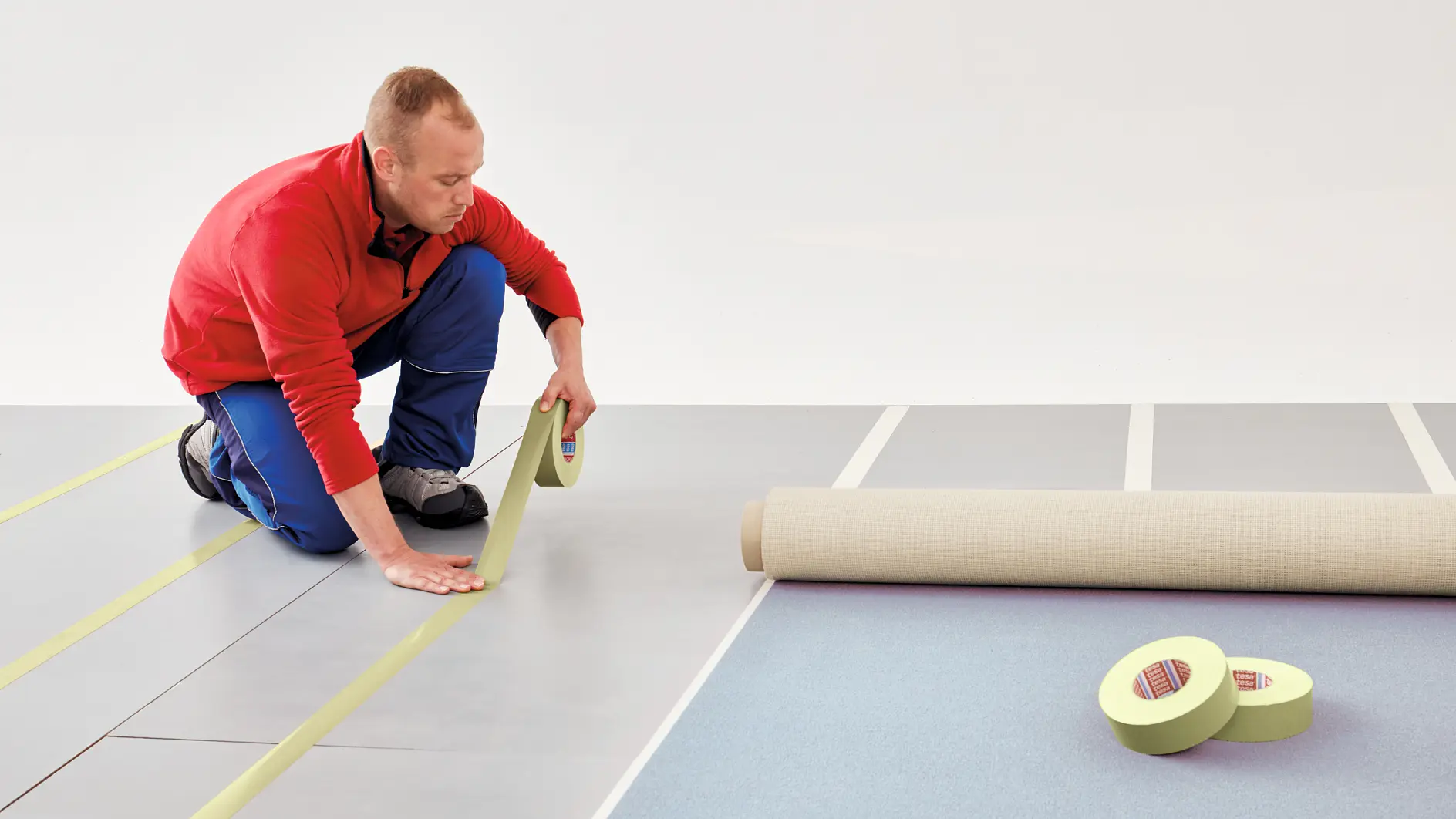 A person in a red jacket and blue pants kneels on a floor, unrolling tesa tape in straight lines. Nearby, there is a rolled-up mat and additional rolls of tesa tape. The background is plain white. (This text has been generated by AI)