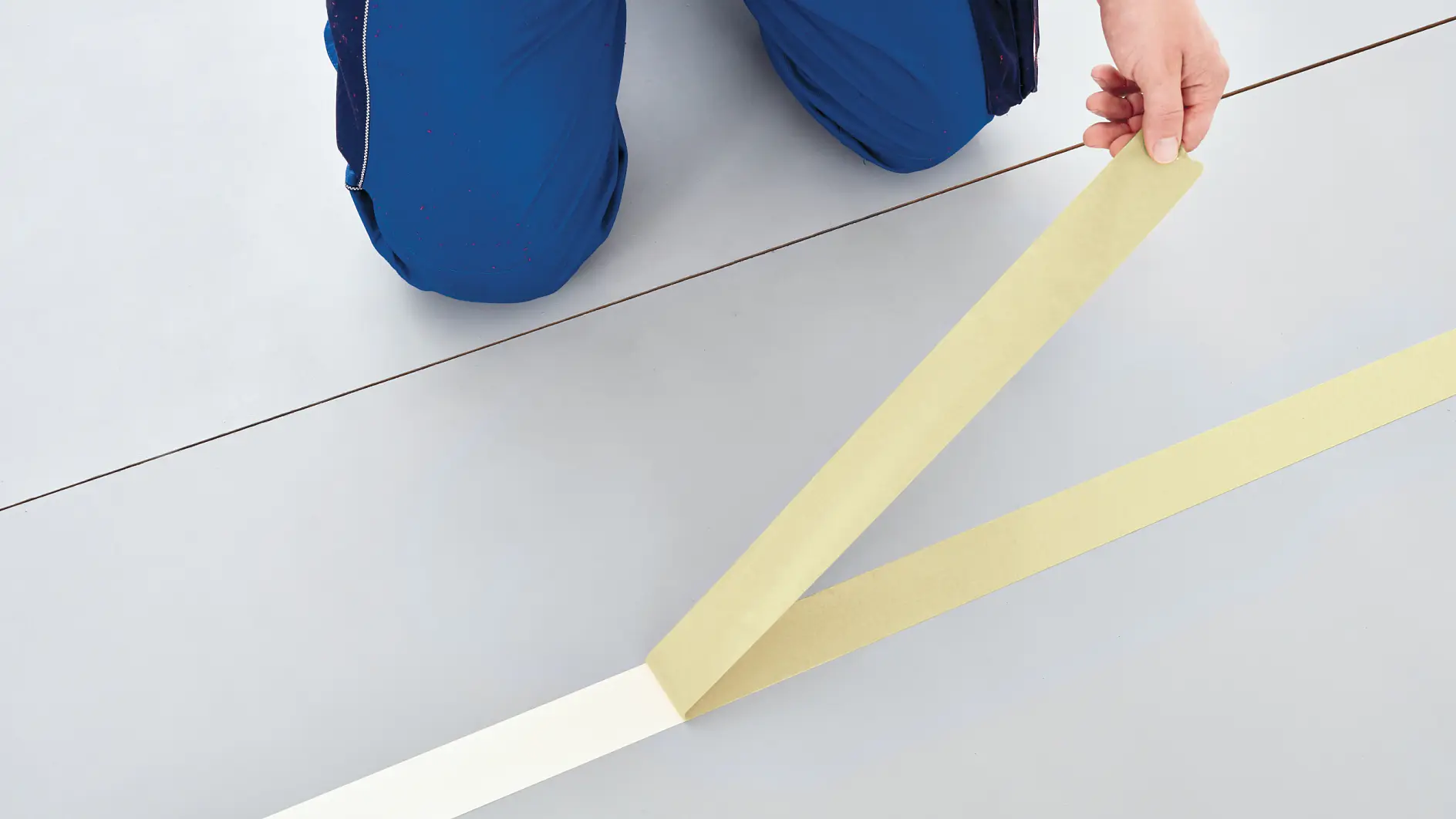 A person in blue pants is kneeling on the floor and applying beige tesa tape in a straight line. The floor is light gray. The persons upper body is not visible in the image. (This text has been generated by AI)