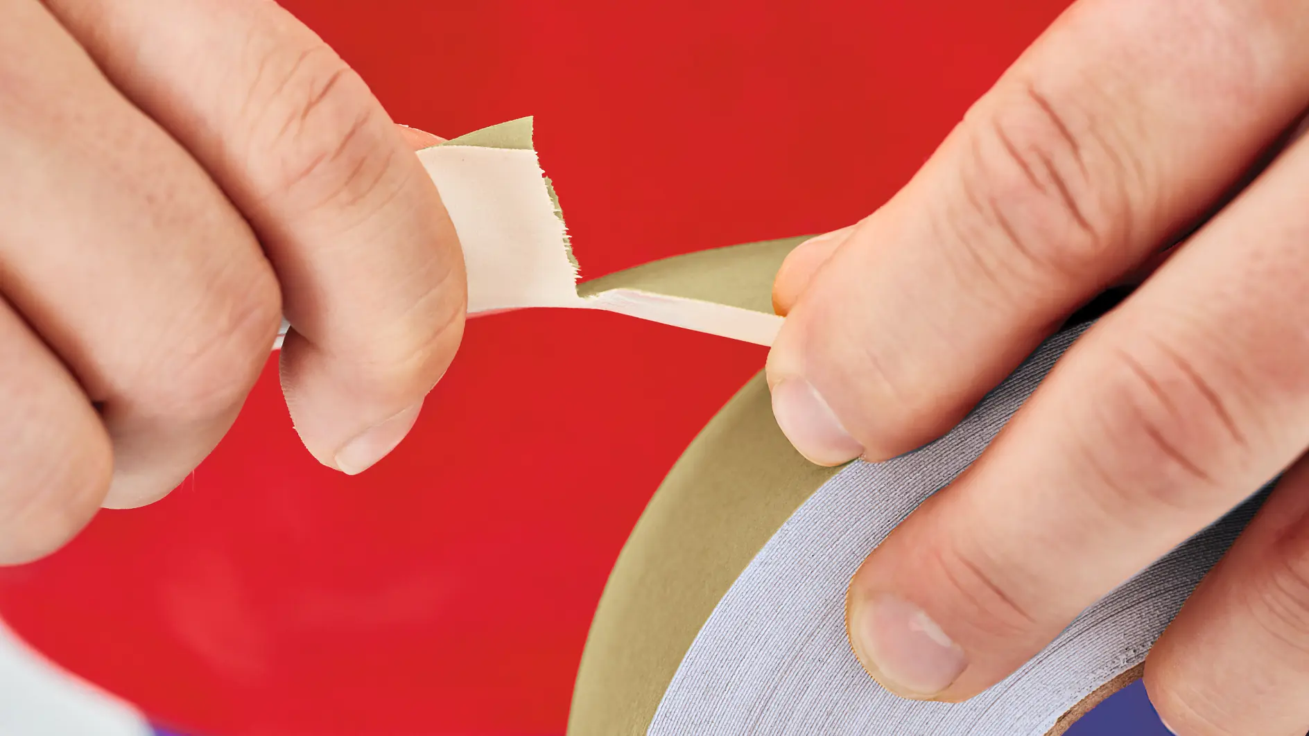 Close-up of hands peeling off a strip of tesa tape from a roll. The background is a vibrant red. The tesa tape appears to be masking or painters tape, usually used for protecting surfaces during painting or crafting tasks. (This text has been generated by AI)