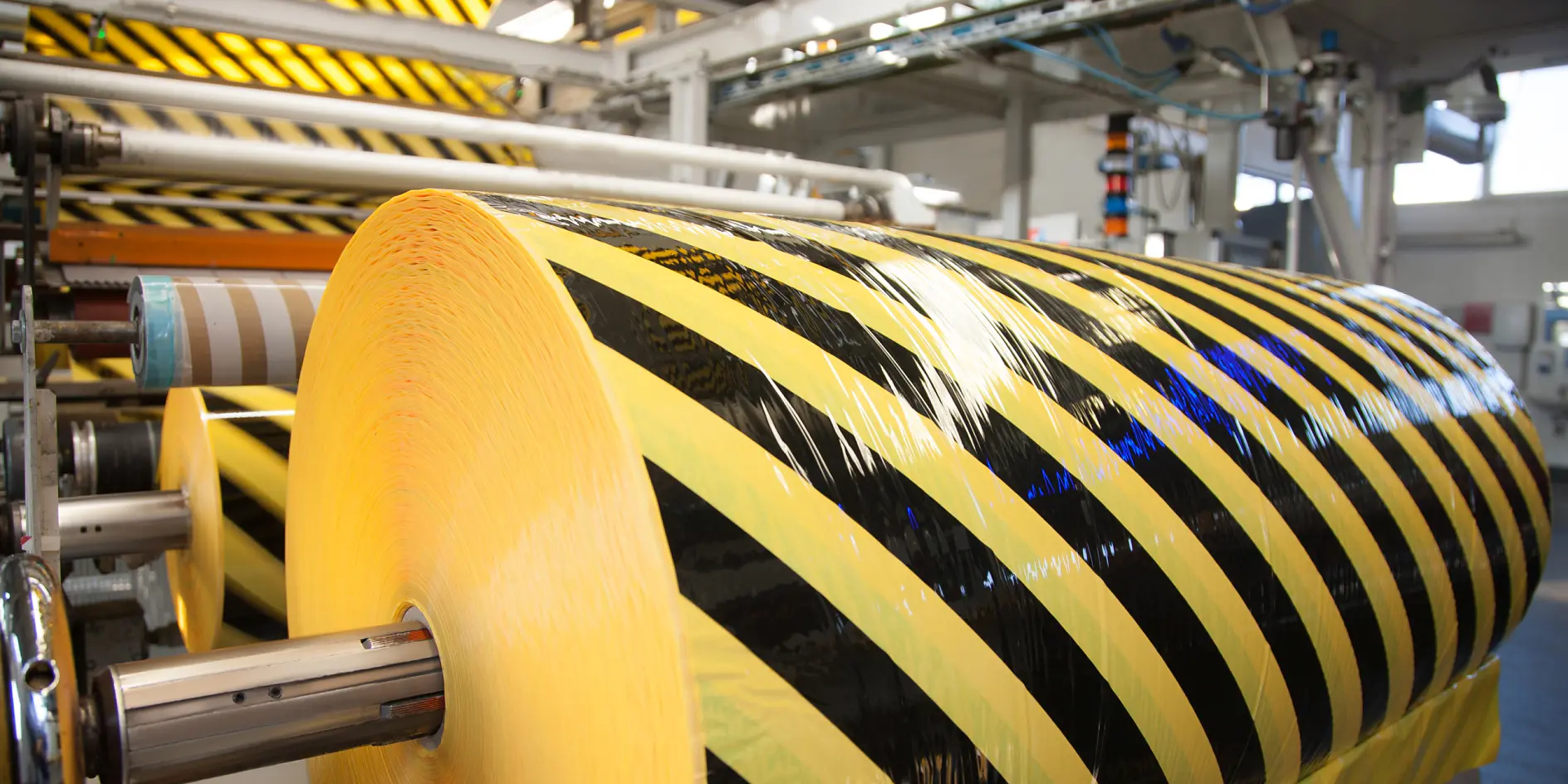 Large rolls of yellow and black caution tesa tape are mounted on metal rods in an industrial setting. Machinery and pipes are visible in the background, suggesting a manufacturing process. The tesa tape features diagonal stripes.