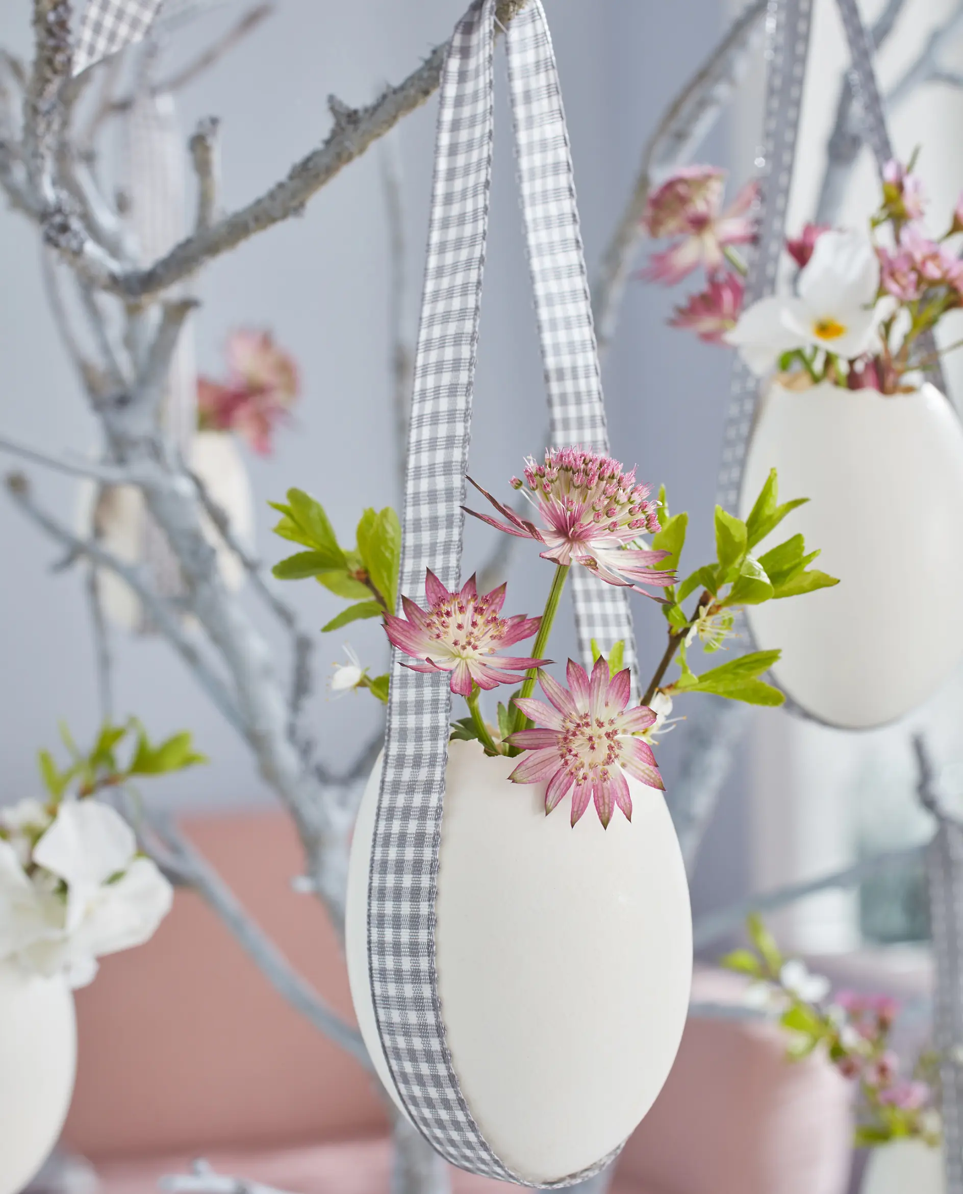 A decorative egg-shaped vase hangs from a branch using a checkered ribbon and is secured with tesa tape. The vase contains pink flowers and green leaves. It is one of several similar vases arranged in the background, with a soft focus on a pink couch partially visible.