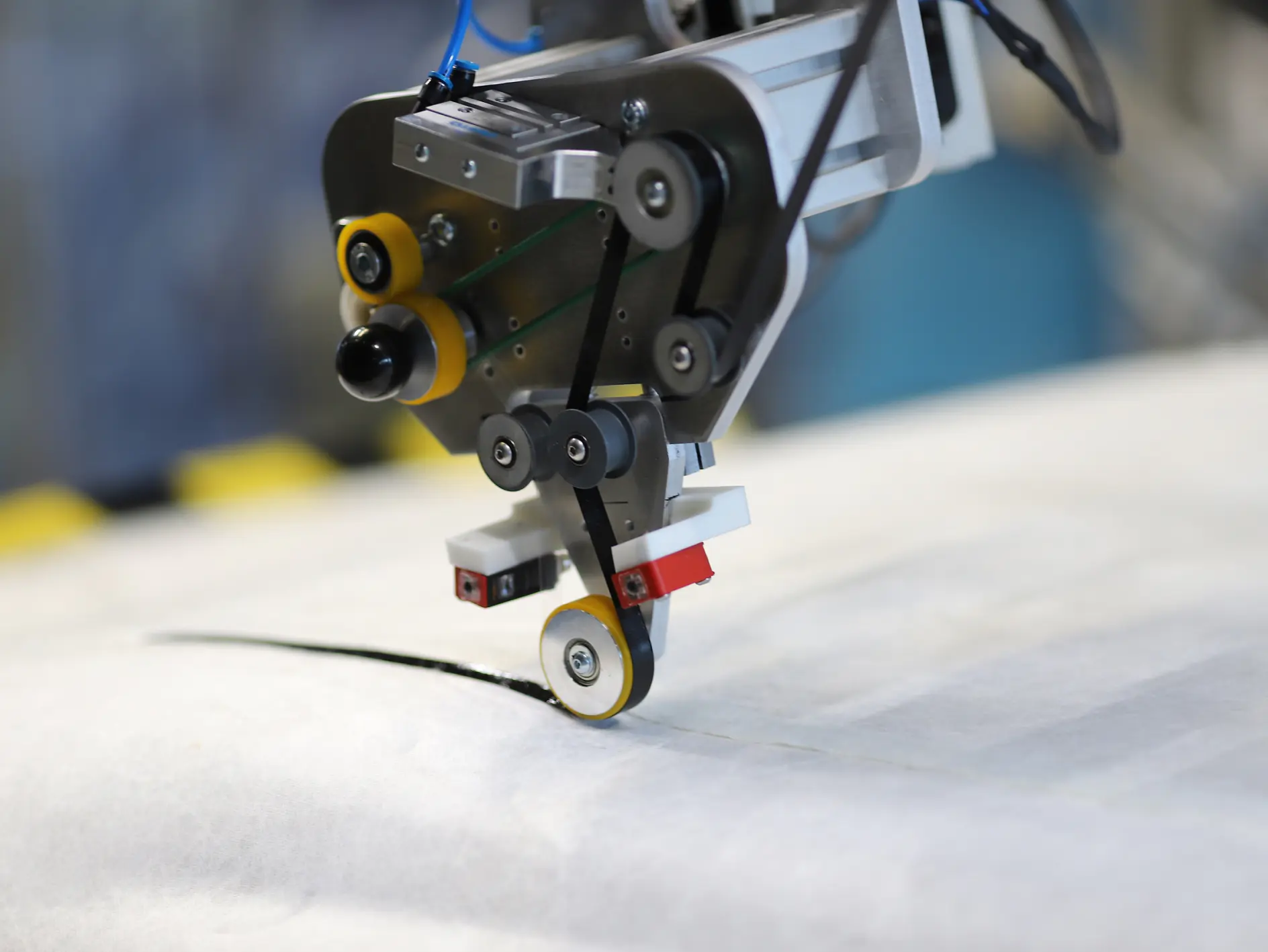 A robotic machine is working on a textile material, using circular cutting blades to make a precise cut. The machinery is equipped with various components and wiring, set against a blurred industrial background. To ensure secure connections and fastenings, tesa tape is used throughout the equipment assembly. (This text has been generated by AI)