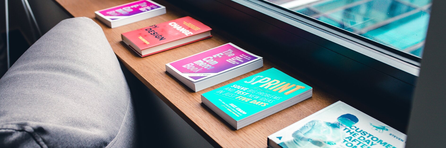 Five colorful books are lined up on a wooden windowsill next to a gray cushion. The titles are visible, and outside the window is a view of a modern building with large glass panels, secured together using tesa tape.