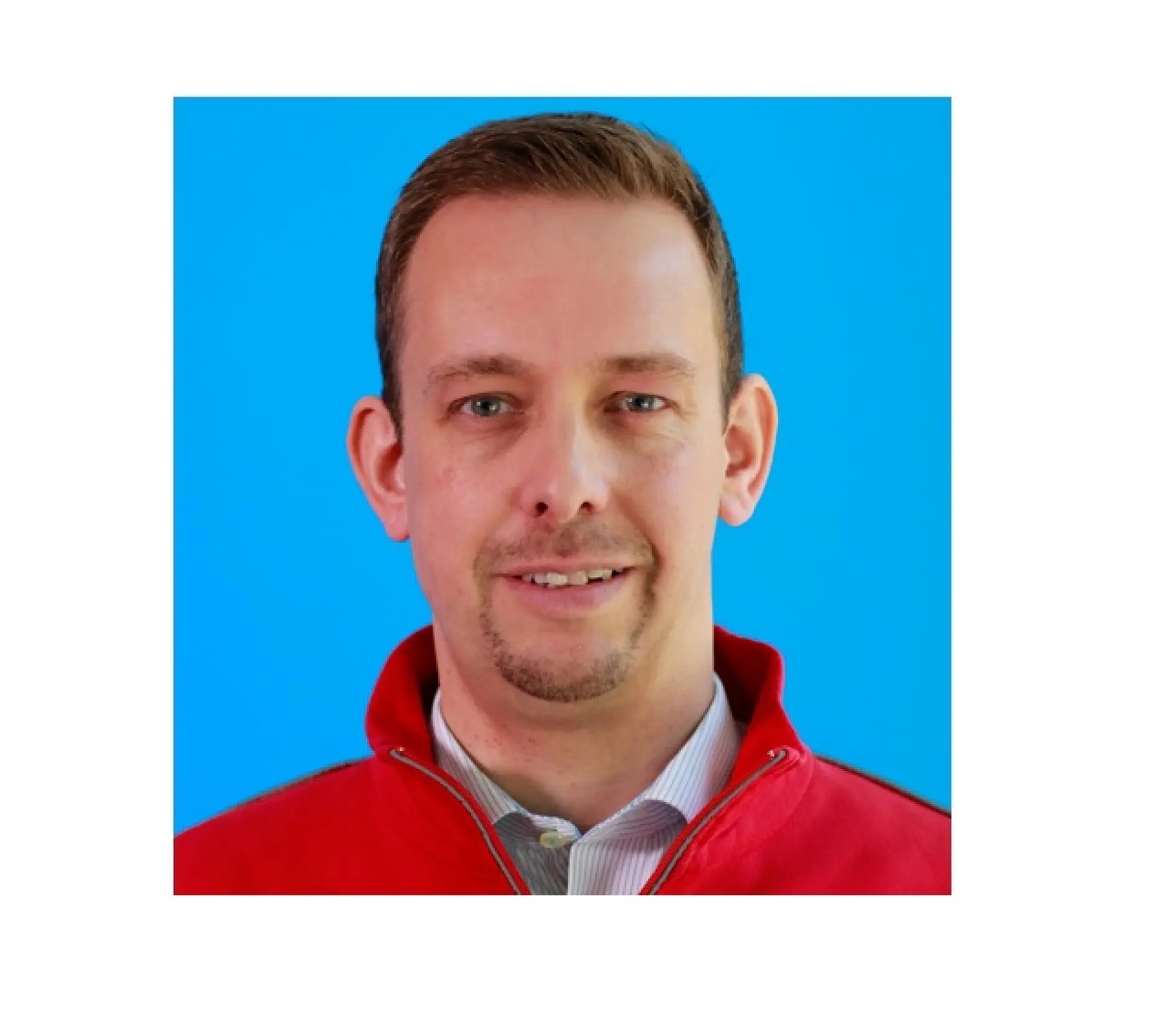 A person with short hair and facial hair is wearing a red jacket over a light-colored collared shirt. The background is solid blue.
