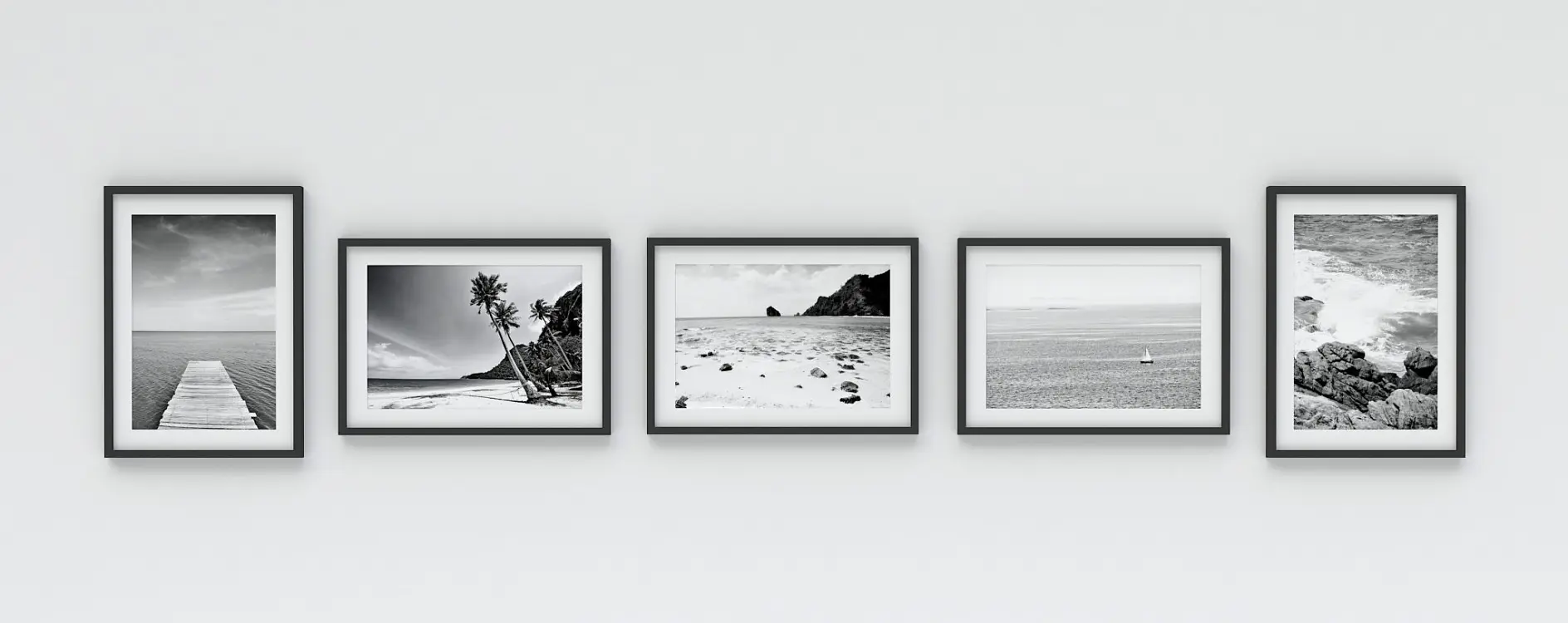 Five black and white landscape photographs are framed in black and arranged in a row on a white wall. The images depict ocean scenes, including a pier, palm trees on a beach, a rocky coast, and expansive seas. These photos are secured with tesa tape for a clean and professional presentation.