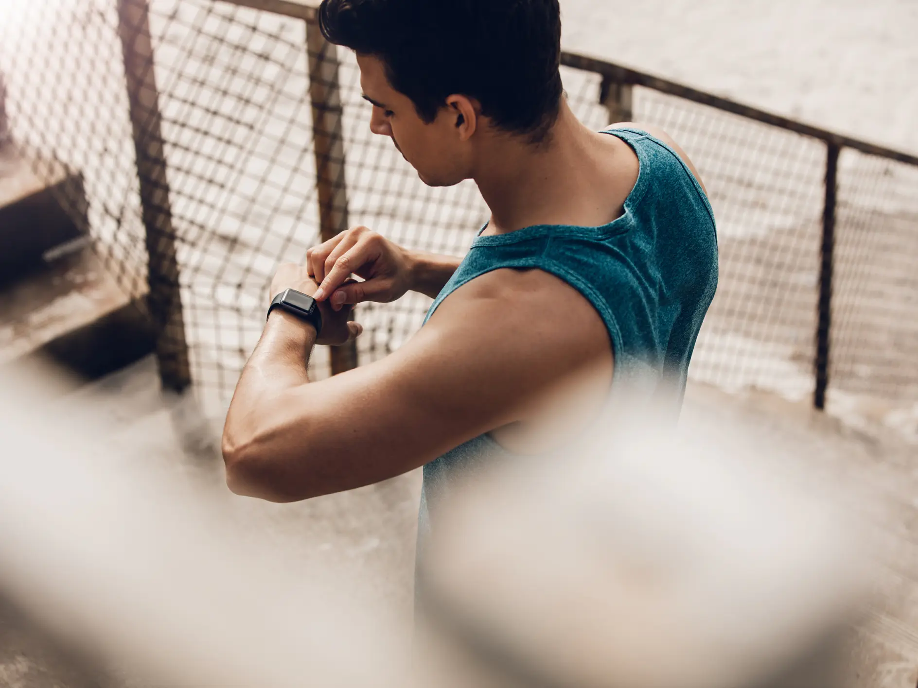 A man wearing a blue sleeveless shirt stands on an outdoor staircase, viewing a smartwatch on his left wrist. The scene is framed by a metal fence, and the ground appears concrete. (This text has been generated by AI)