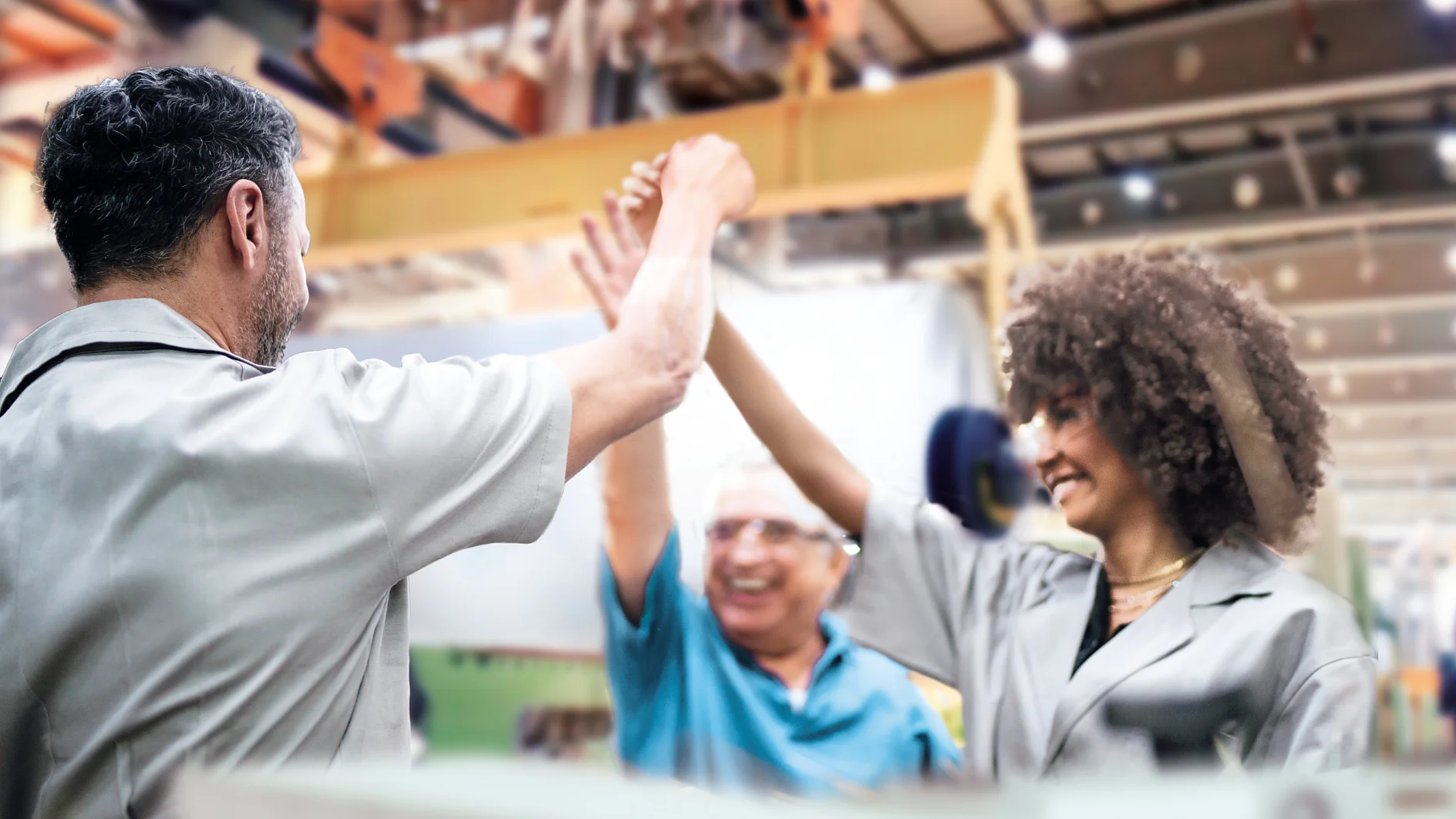 Three factory workers in uniforms celebrate inside an industrial setting, smiling and giving a high-five. The background shows machinery and bright lighting, along with rolls of tesa tape prominently displayed on the shelves. (This text has been generated by AI)
