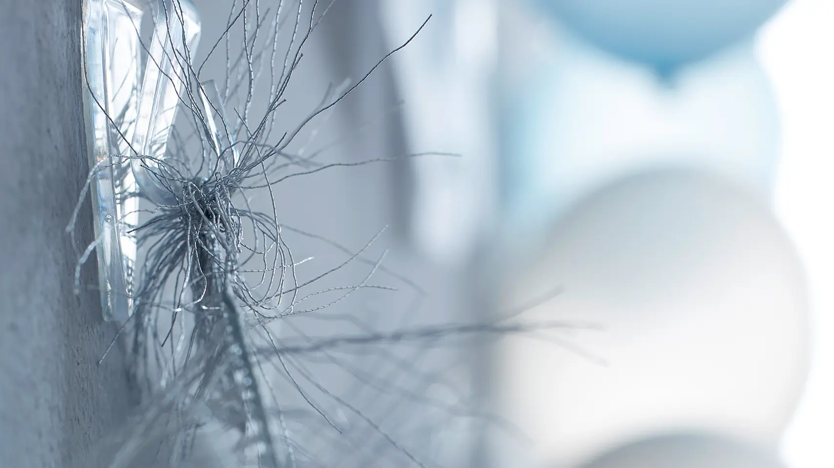 Close-up of metallic tinsel hanging on a gray wall with adhesive hooks. In the blurred background, there are blue and white balloons. The focus is on the tinsel, and the scene suggests a festive decoration secured with tesa tape.