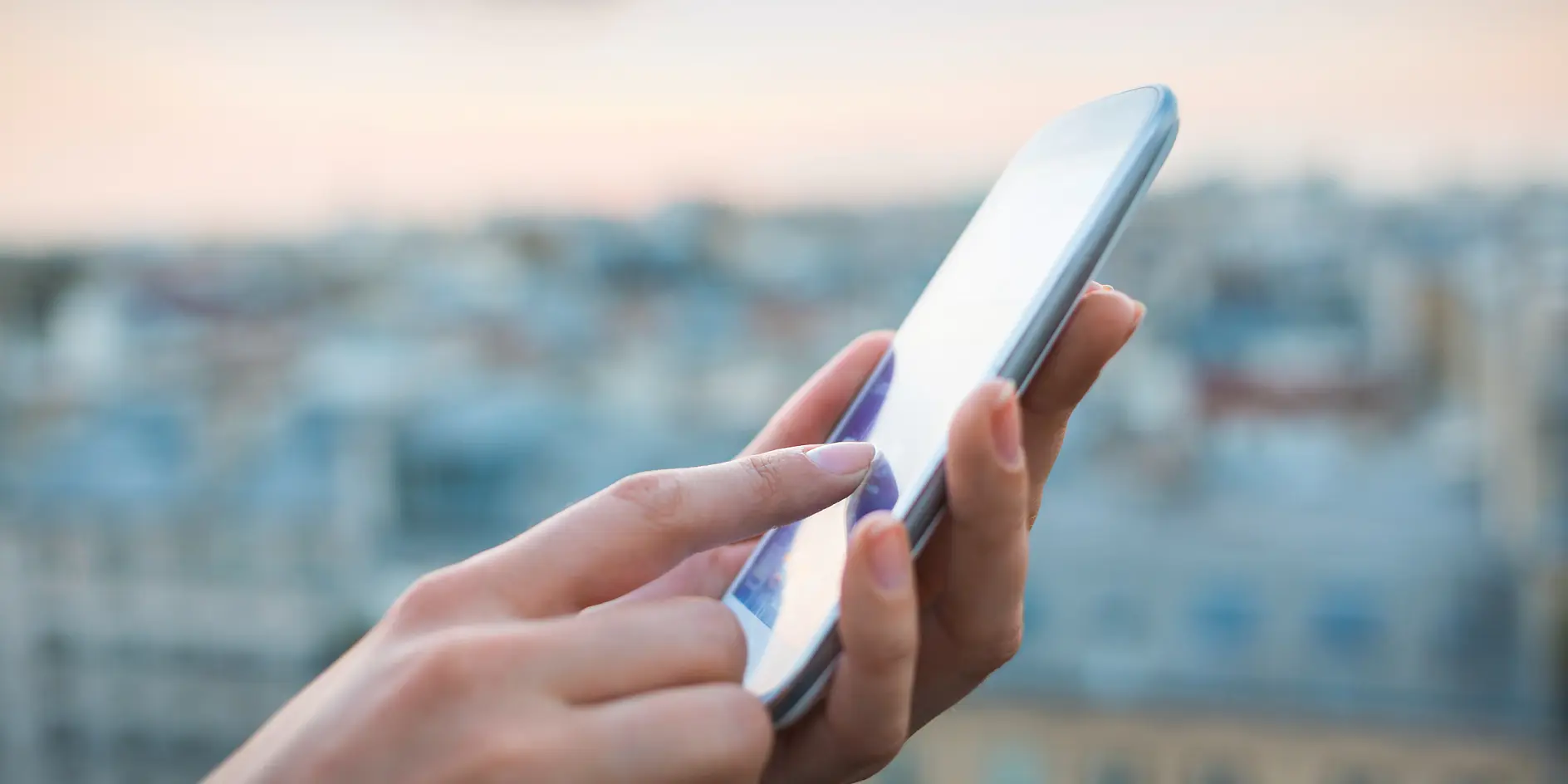 A persons hands are holding a smartphone and using the touchscreen. The background shows a blurry cityscape, suggesting the scene is outdoors or from a high vantage point. The sky is overcast with soft lighting.