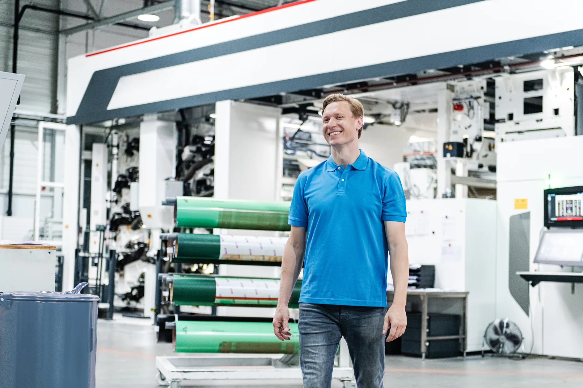 A man in a blue polo shirt and jeans is standing in a large industrial facility. Behind him, there is machinery and large rolls of tesa tape material. The setting appears to be a printing or manufacturing plant. (This text has been generated by AI)