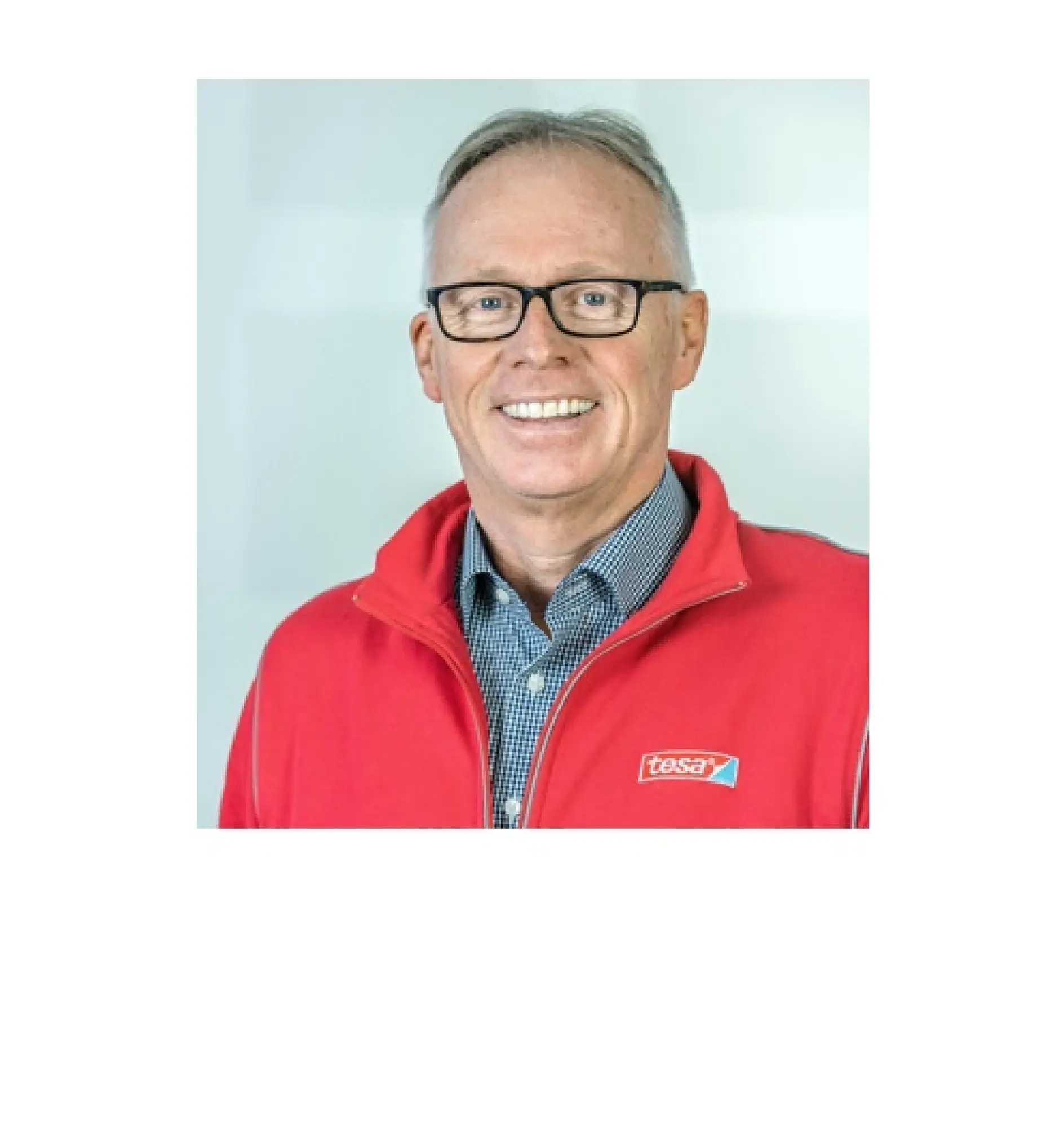 A person wearing glasses and a red jacket with a tesa logo is smiling. They have short hair and are standing against a plain background.