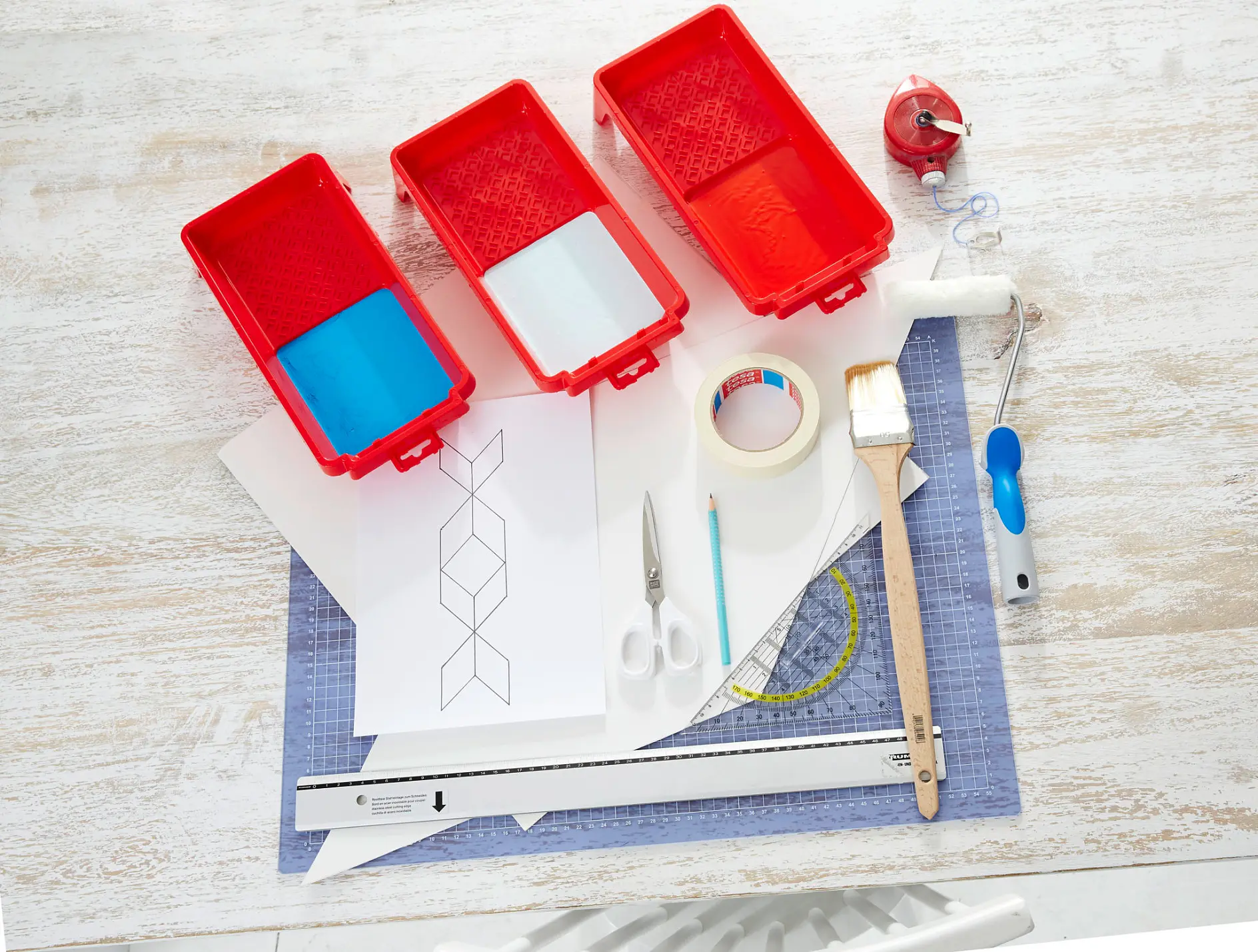 A workspace with three red paint trays, tesa tape, a brush, scissors, a cutter, and a ruler. White paper with a geometric design is laid out on a cutting mat. A tape measure and a paint roller are also visible on the wooden surface.