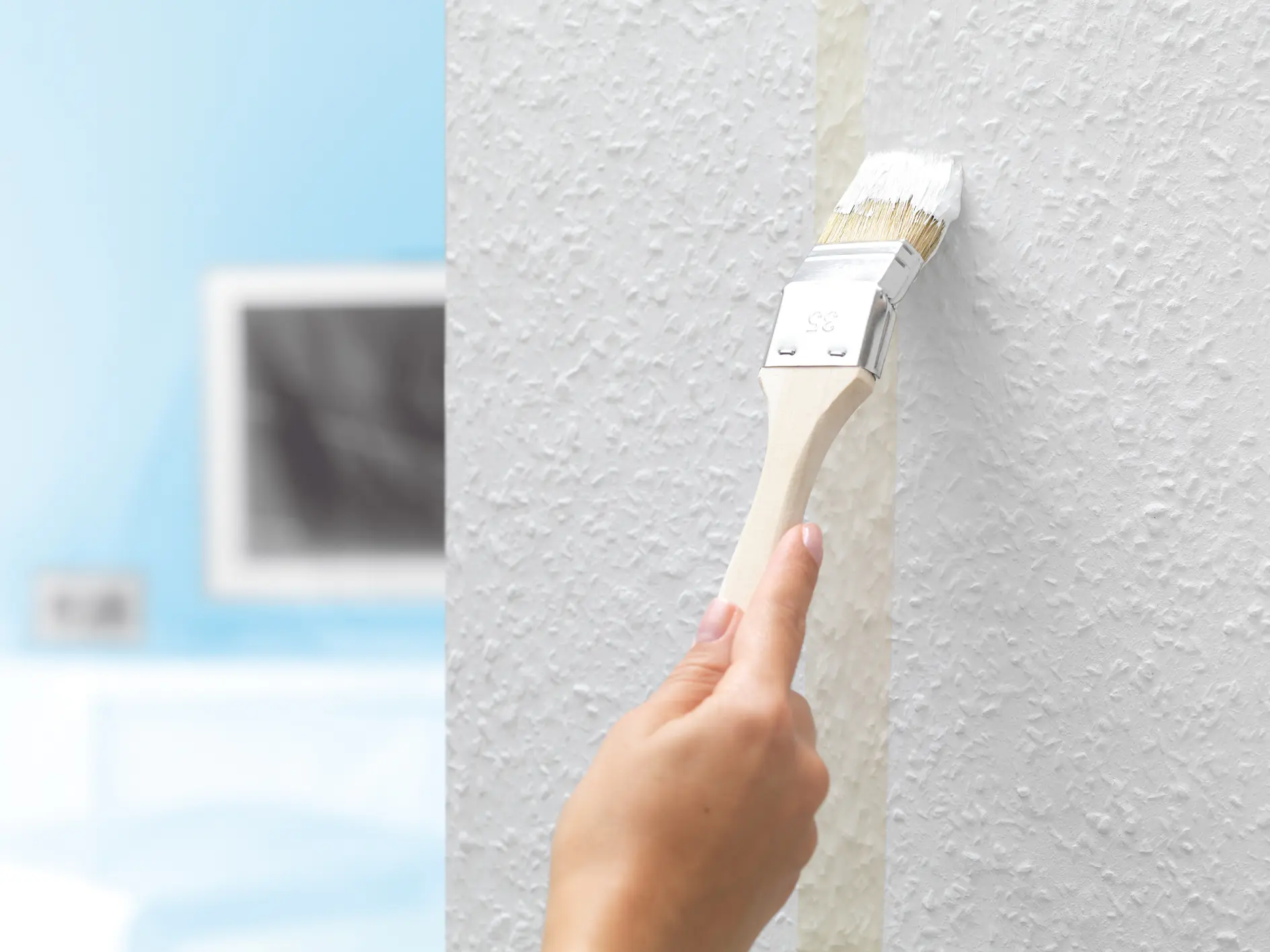 A hand holding a paintbrush applies white paint neatly along the edge of a textured wall with tesa tape. The background shows a blurred room with a light blue wall and a television.