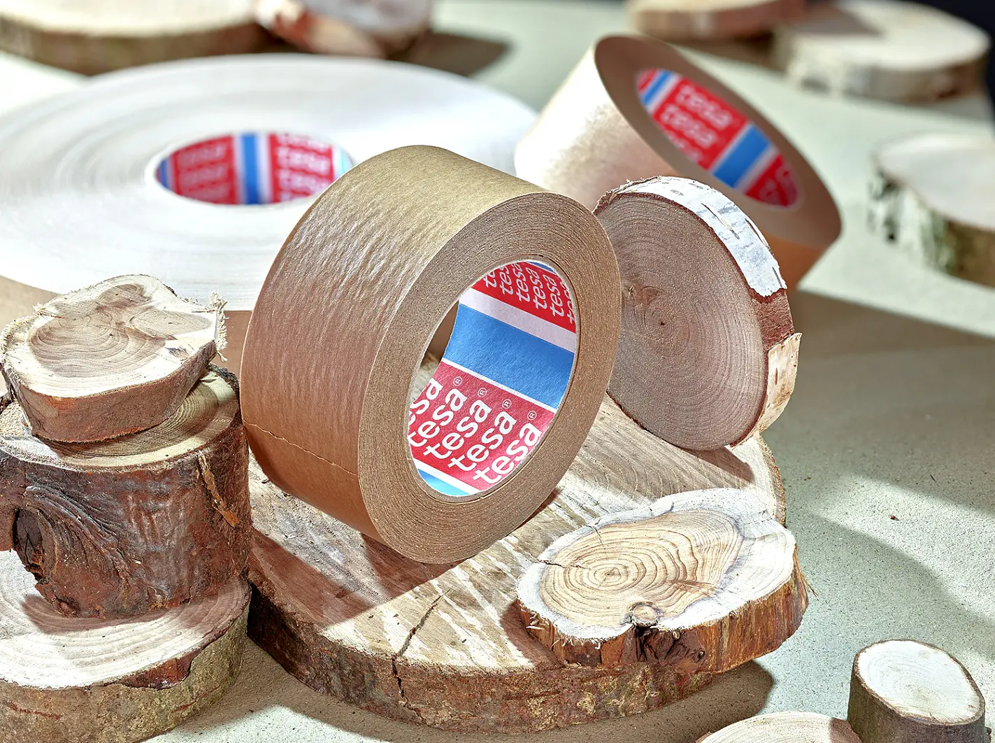 A roll of brown tesa tape is placed on round wooden slices. The tesa tape features a red and blue label inside. The wooden slices vary in size and have visible tree rings, adding a natural element to the setup. (This text has been generated by AI)