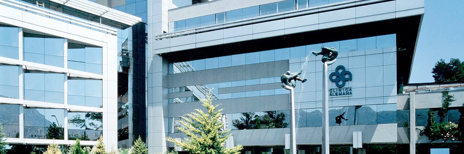 Modern glass and concrete office building with reflective windows and angular design. The foreground features a landscaped area with shrubs and trees. A logo is visible on the building facade, made prominent by its use of tesa tape. Clear blue sky above.