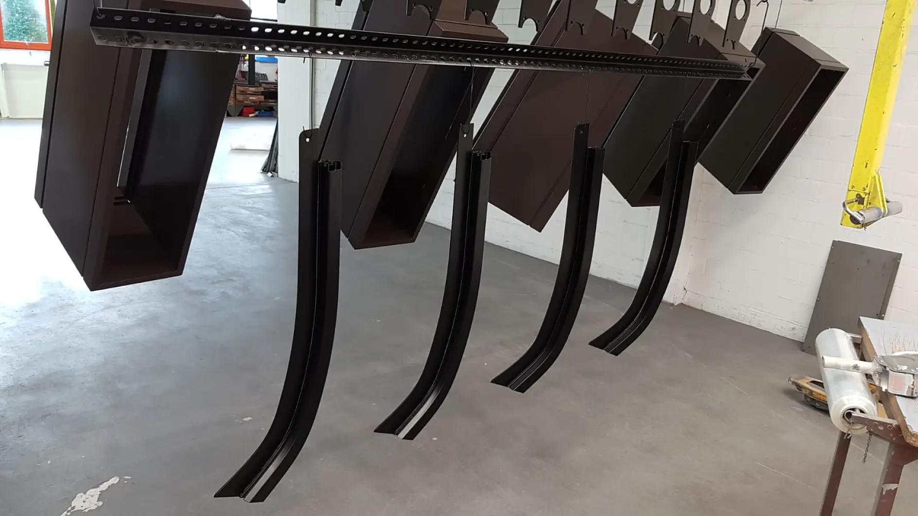 Several metal components, including rectangular boxes and curved bars, are hanging from hooks attached to a metal frame in what appears to be a workshop or assembly area with a concrete floor and white brick walls. (This text has been generated by AI)