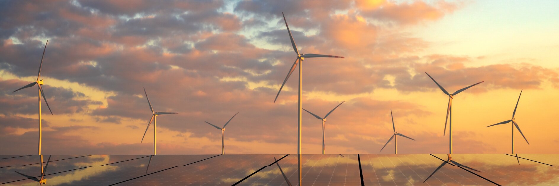 Solar panels with reflections of the sky cover the ground, while several wind turbines stand behind them. The sky is partly cloudy with a warm glow from the setting or rising sun.