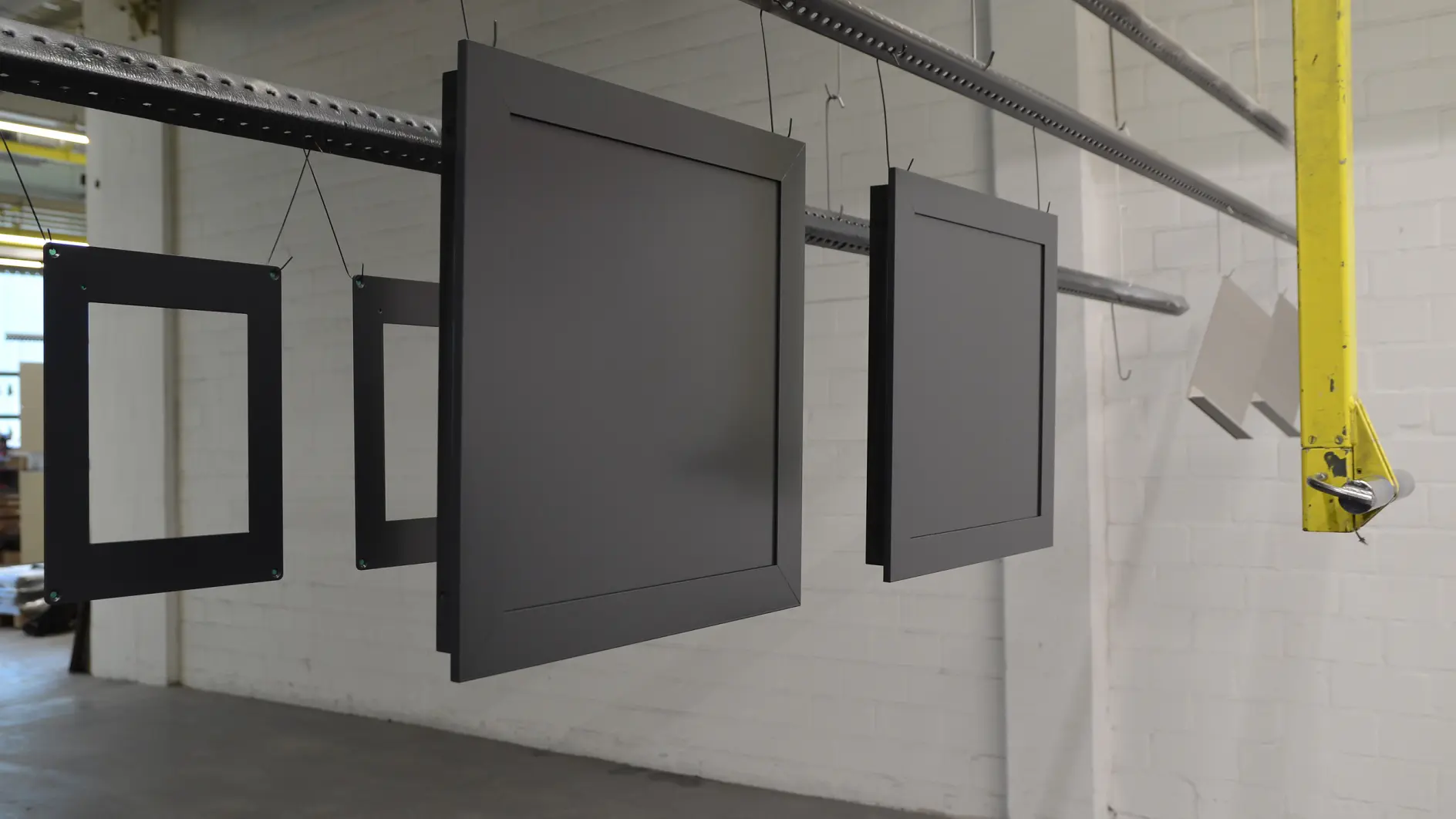 A series of rectangular frames hang from a metal rod in an industrial setting. The frames are painted dark gray and are suspended in a row against a background of white brick walls and a concrete floor, all secured using tesa tape. (This text has been generated by AI)