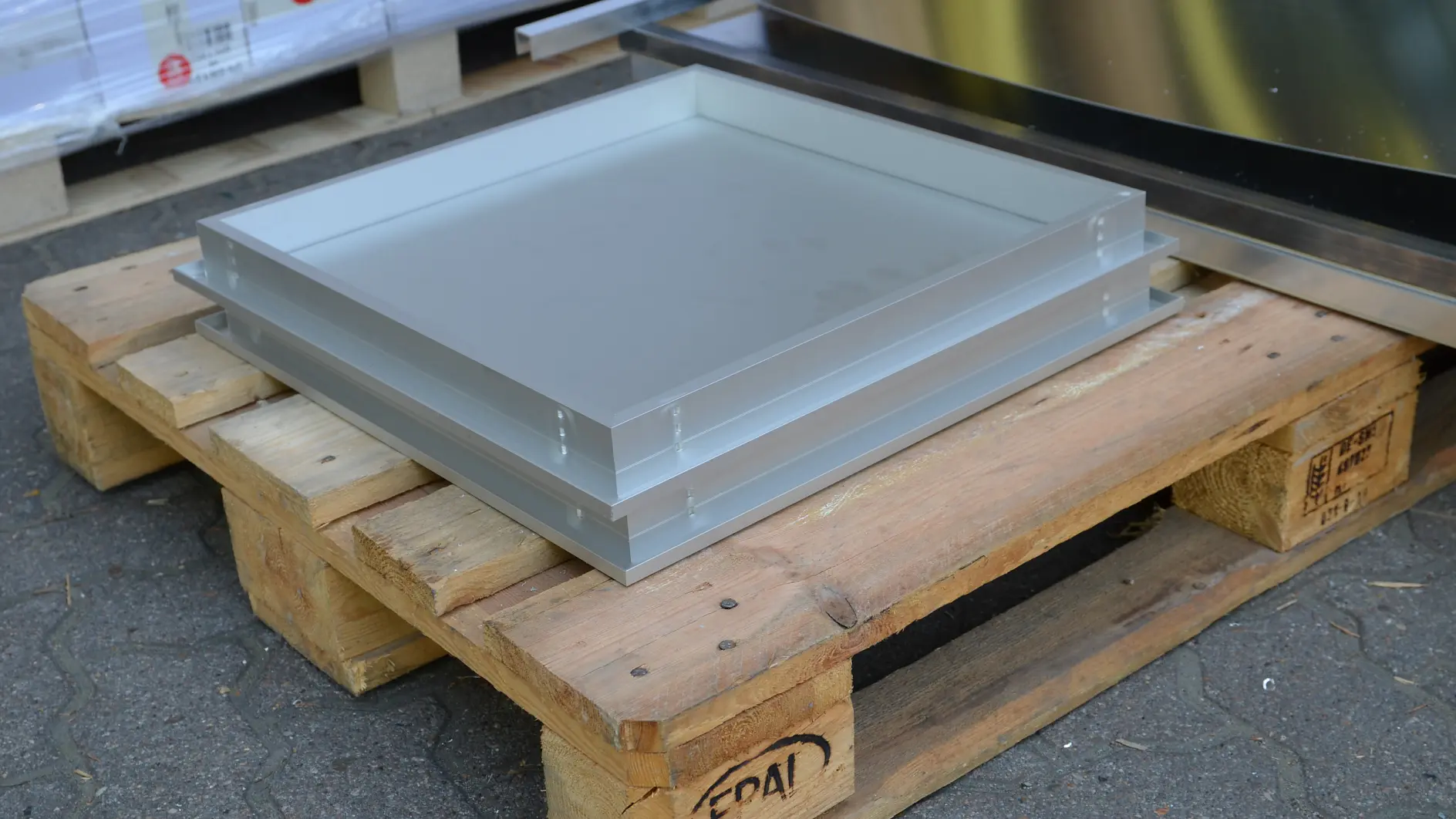 A metal tray with a shallow edge is secured using tesa tape on a wooden pallet. The pallet is situated on a paved outdoor surface. Another reflective metal sheet or tray, also secured with tesa tape, is partially visible in the background on the pallet. (This text has been generated by AI)