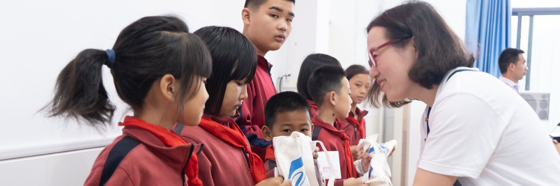 A woman distributes gift bags to a group of children in a classroom setting. The children are standing in a row, wearing red and black uniforms with red scarves. The woman is using tesa tape to secure the wrapping on each gift bag. She is wearing glasses and dressed casually.