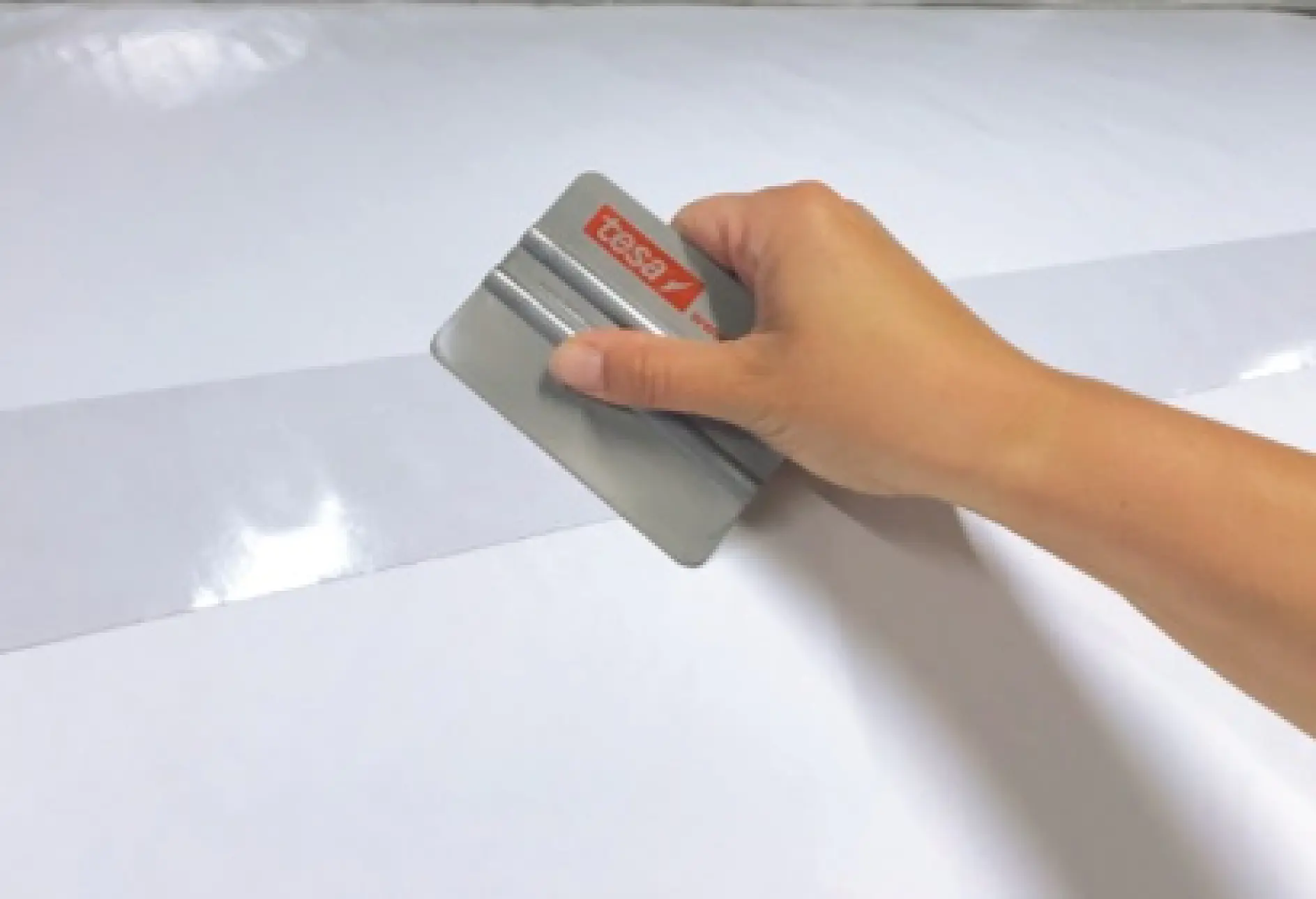 A hand holding a tesa branded scraper is smoothing or applying adhesive to a flat surface with a visible shiny strip on it. The background is plain and light-colored. (This text has been generated by AI)
