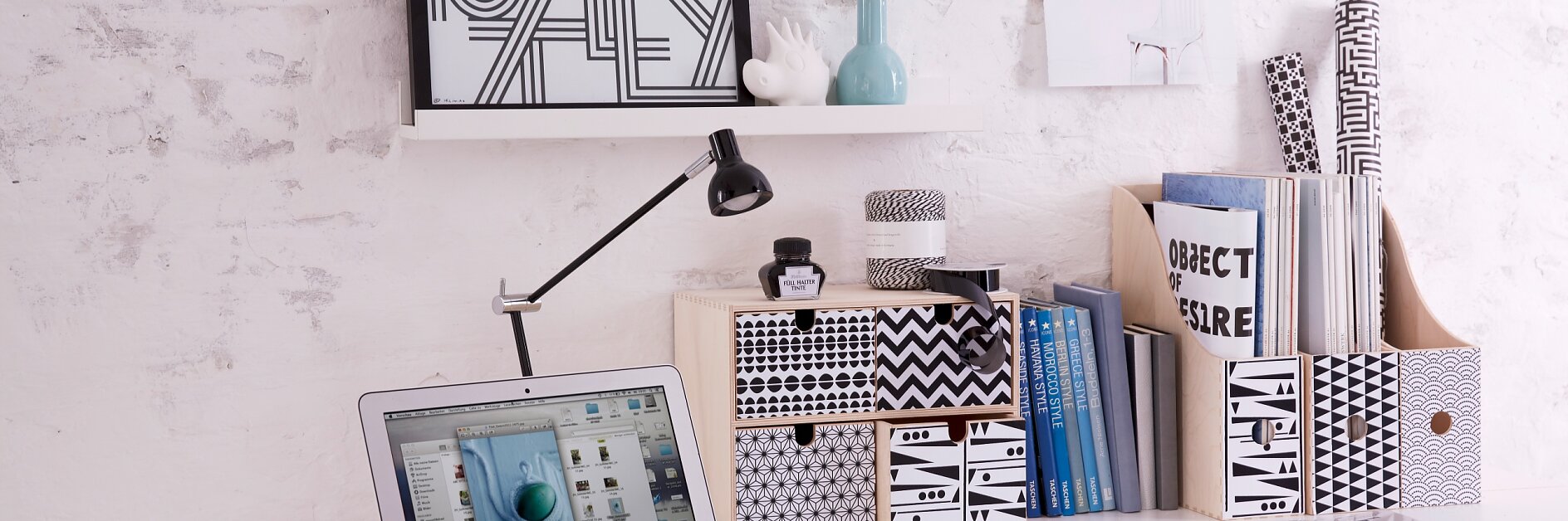 A minimalist white desk with a laptop displaying an image. Above are shelves with decorative boxes, books, and small vases, along with a framed print and a magazine page fastened using tesa tape. A black desk lamp is positioned on the desk.