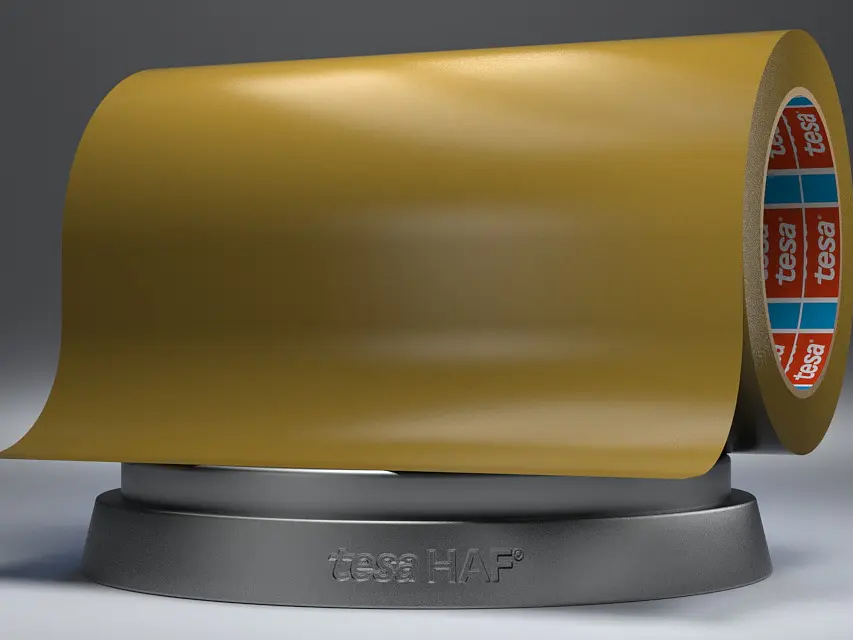 A large roll of yellow adhesive tesa tape is displayed on a gray base against a dark background. The inner side of the tesa tape roll has a red, blue, and white pattern.
