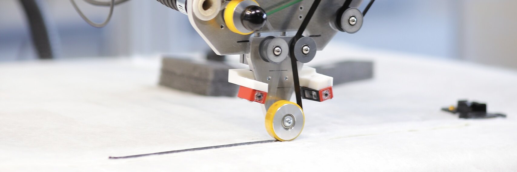 Close-up of an industrial machine component with a large black spool and metallic parts. The setup includes various cables, gears, and a mechanism feeding material through the tesa tape. The background is blurred, suggesting an industrial environment.