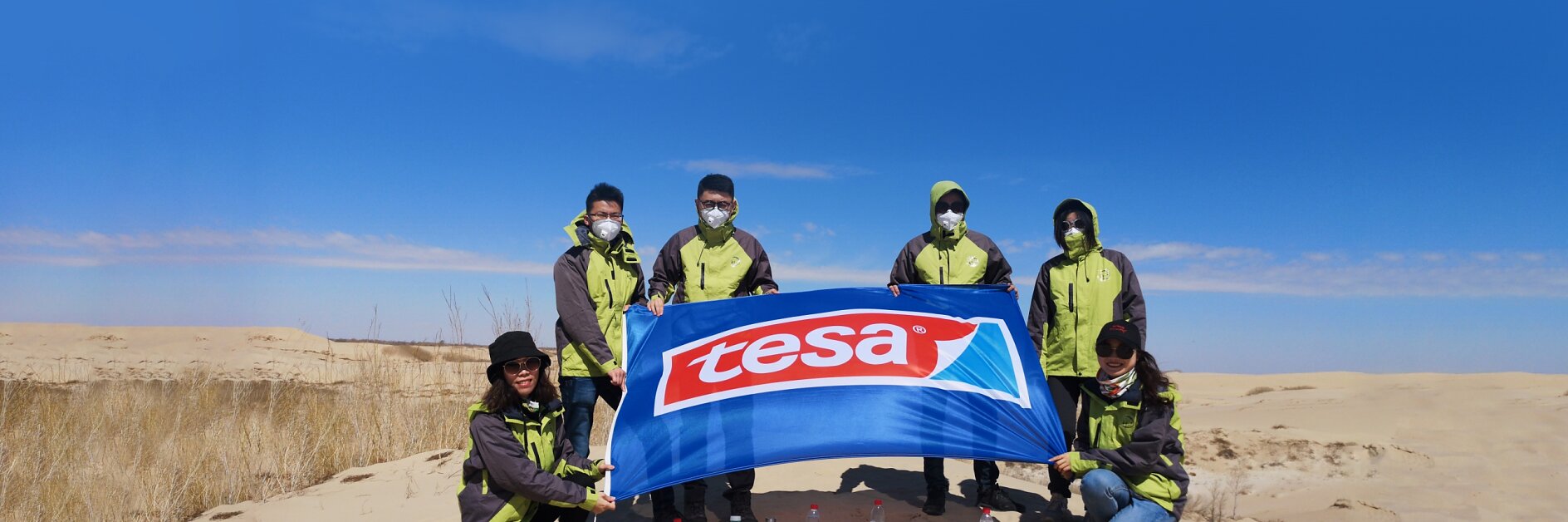 Six people wearing green jackets and masks stand on a sandy landscape, holding a blue flag with the red and white tesa tape logo. They are in a desert-like area with a clear blue sky and sparse vegetation. Several water bottles are placed on the sand.