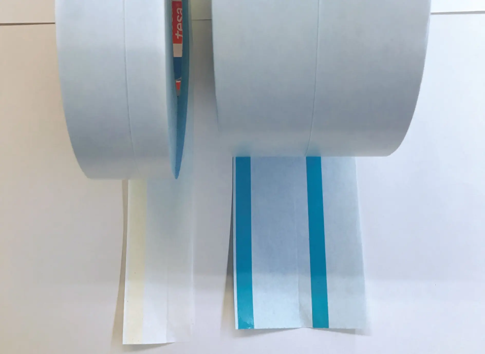 Two rolls of white masking tesa tape, partially unrolled and placed on a flat surface. The left tesa tape has a plain strip, while the right tesa tape has a blue line running through the center. (This text has been generated by AI)
