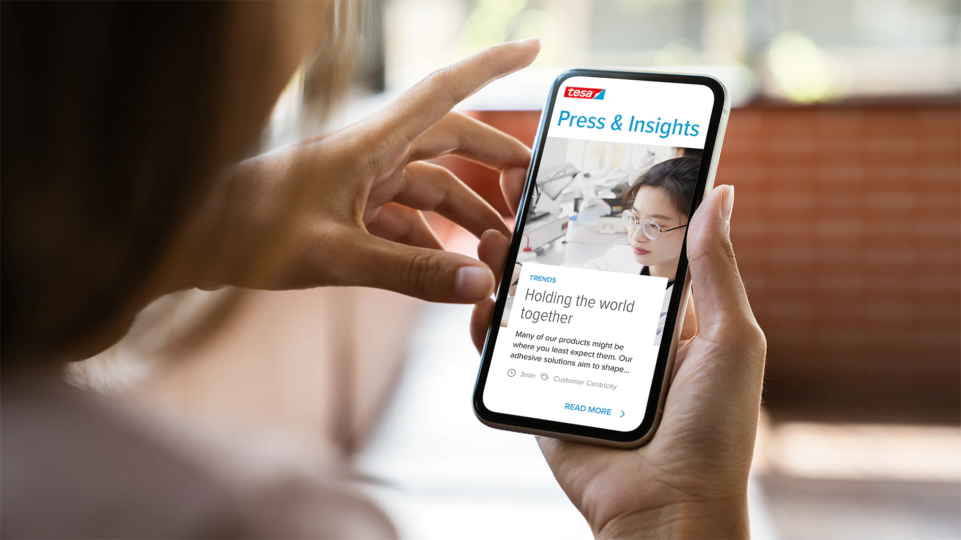 A person holding a smartphone displaying a webpage titled Press & Insights. The screen shows an article with an image of a woman behind a microscope and text about tesa tape adhesives. The smartphone is held in a hand with a blurred background. (This text has been generated by AI)