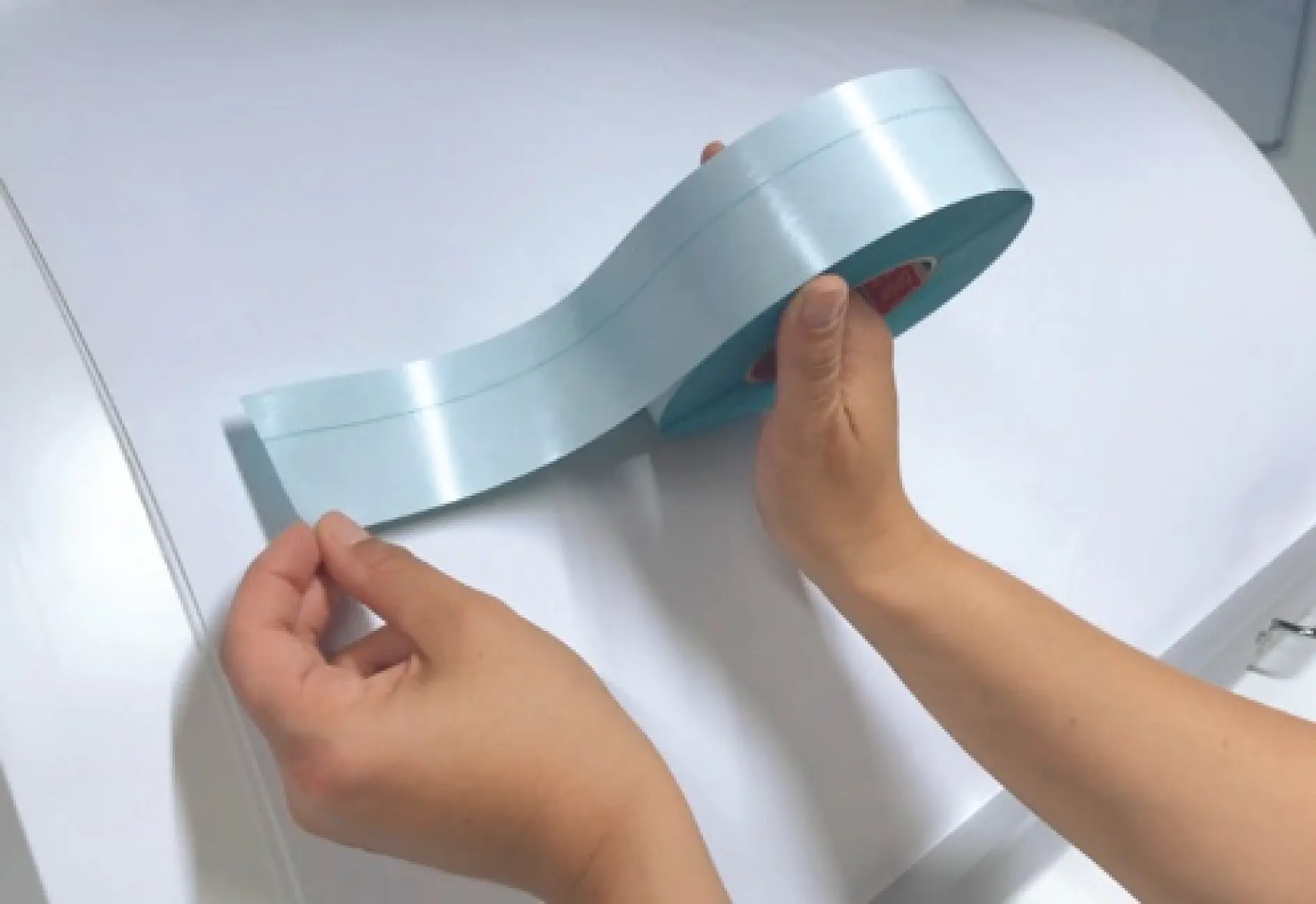 Hands holding a roll of light blue tesa tape above a white surface. (This text has been generated by AI)