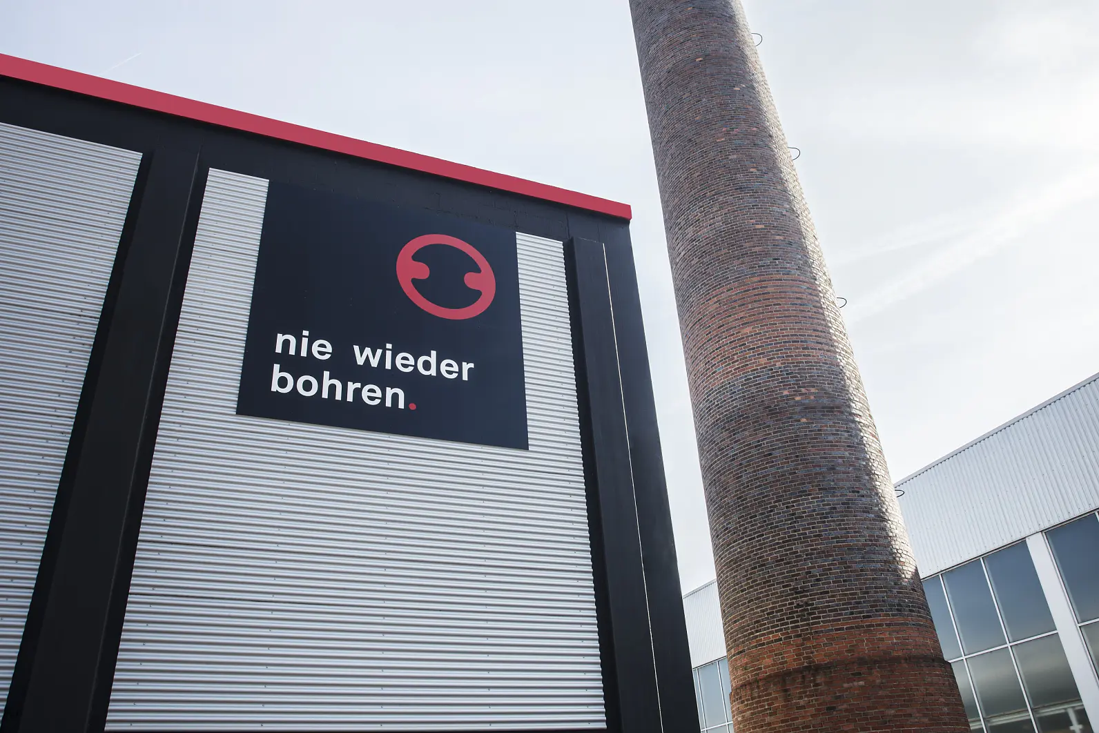 The image shows a building with a sign displaying the text nie wieder bohren. below a circular logo. A tall chimney is visible to the right of the building. In the image foreground, there is tesa tape being used in an installation. The sky is clear with some contrails. (This text has been generated by AI)