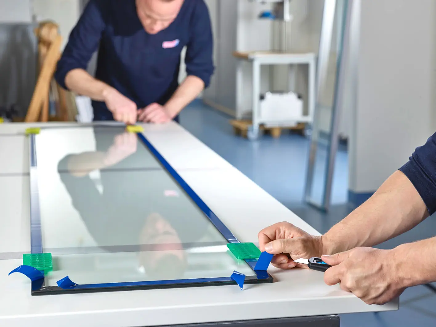 Two people are working in a workshop with a large glass pane on a white table. One person is cutting blue tesa tape, while the other is adjusting the tesa tape on the glass. The background includes various tools and workshop equipment. (This text has been generated by AI)