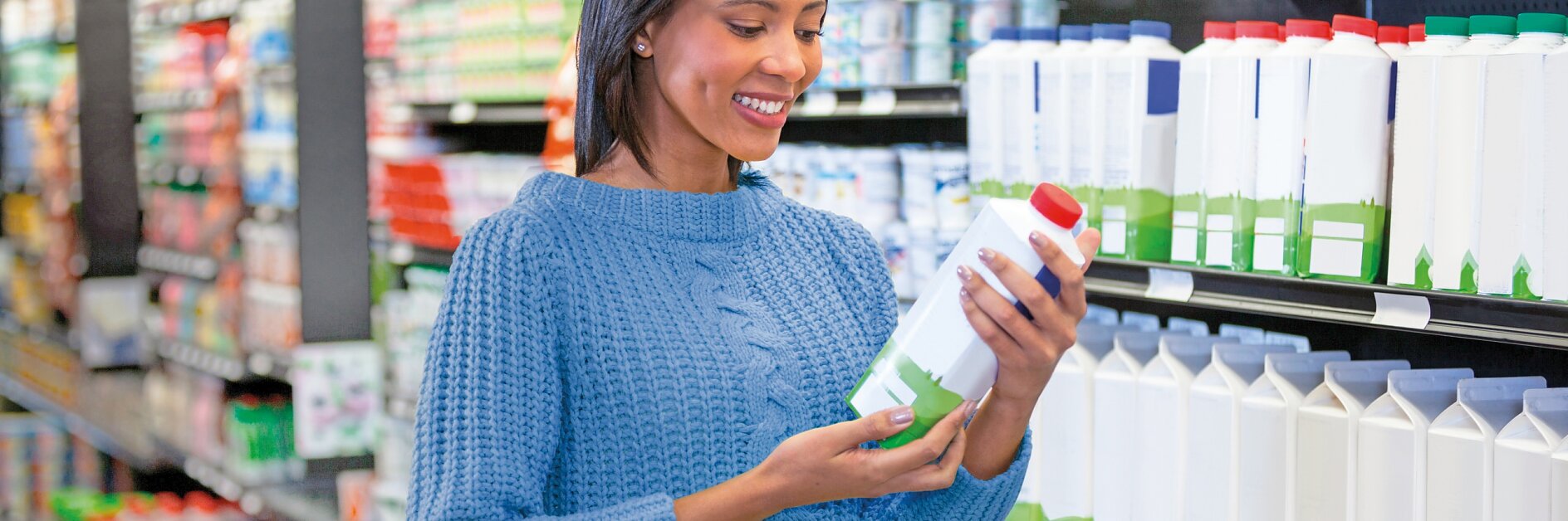 A woman in a blue sweater holds a carton and smiles while standing in a grocery store aisle. She is next to shelves stocked with various bottles and cartons. A red shopping basket with items is beside her.