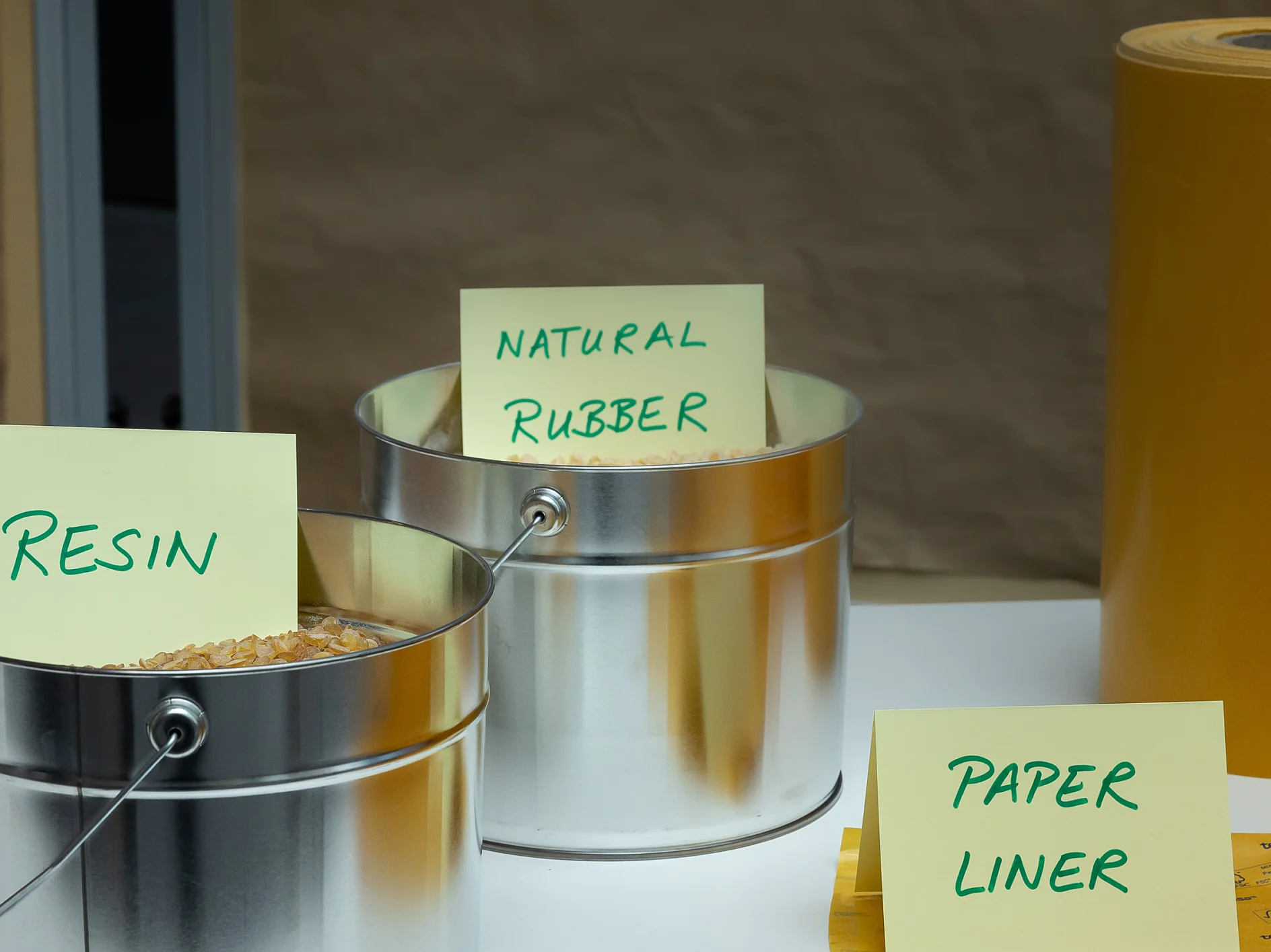 Metal containers labeled Resin and Natural Rubber hold raw materials. A tesa tape Liner sign is placed in front of yellow rolls on a table. (This text has been generated by AI)