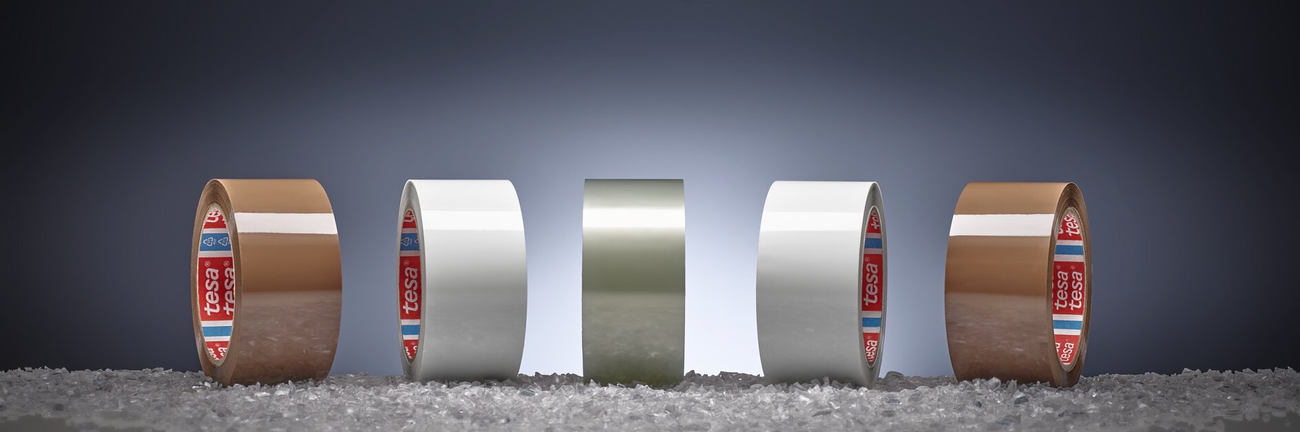 Four rolls of adhesive tesa tape, two brown and two white, are lined up on a surface scattered with small granules. Each roll is standing upright in front of a dark gradient background.