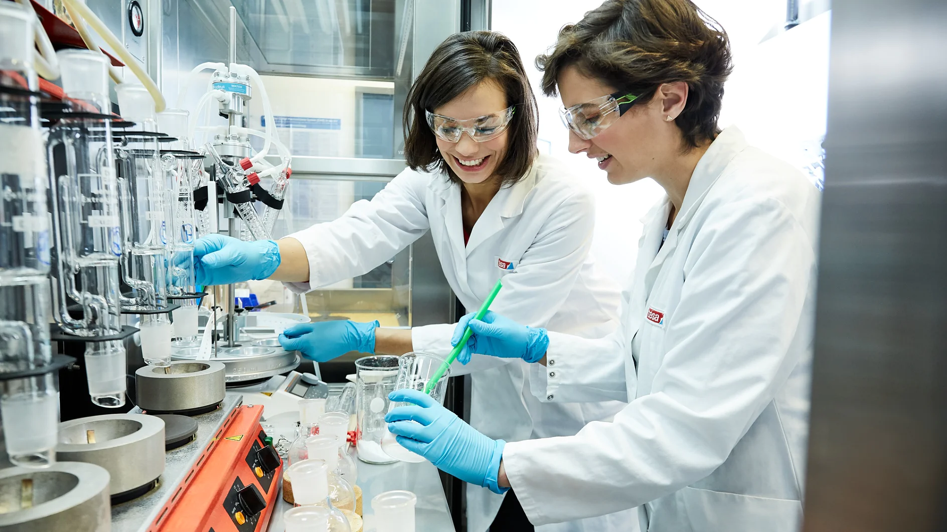 Two scientists in lab coats and safety goggles work at a laboratory bench with various equipment. They are handling chemicals, with one stirring a beaker. Both appear engaged in a research activity, utilizing tesa tape for their experimentations. (This text has been generated by AI)