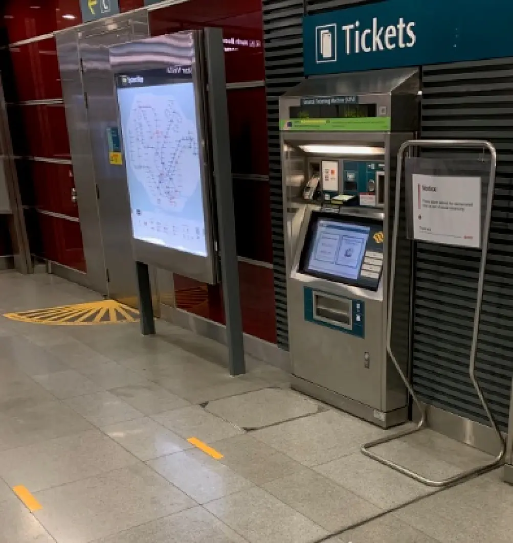 Ticket vending machine at a transit station next to a large map display. A sign is posted on a stand beside the machine, secured using tesa tape. The floor is tiled, with yellow directional arrows. The area is well-lit with metallic and red wall panels. (This text has been generated by AI)