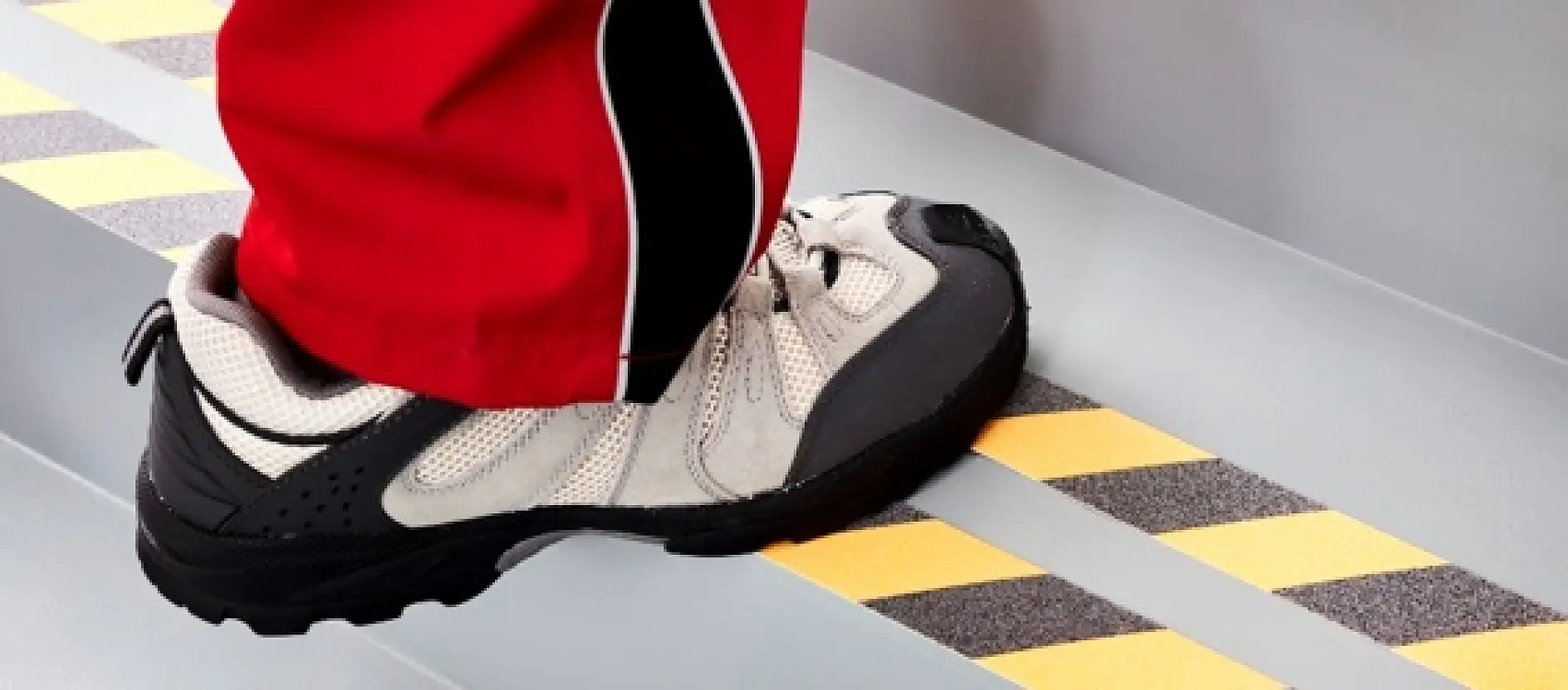 A person wearing a white and gray sneaker with black details steps on a striped black and yellow safety marking made from tesa tape on a gray stair. The leg is clothed in red pants with black and white accents. (This text has been generated by AI)