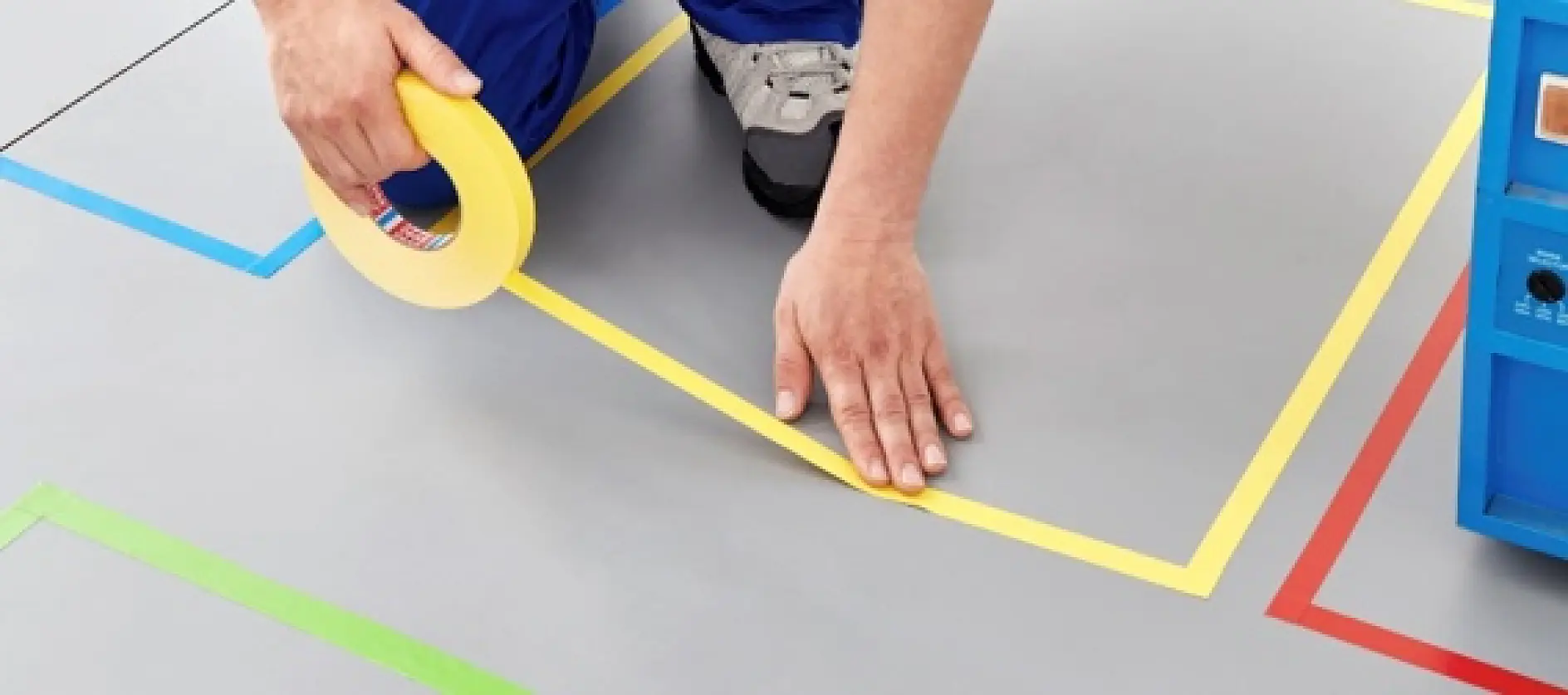 A person wearing blue pants and gray shoes is applying yellow tesa tape on a gray floor within a marked geometric area with colored lines. A blue object is partially visible on the right side of the image. (This text has been generated by AI)
